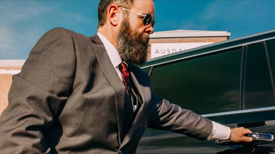 Man in a suit and sunglasses leaning into a car, reaching toward the open door outdoors