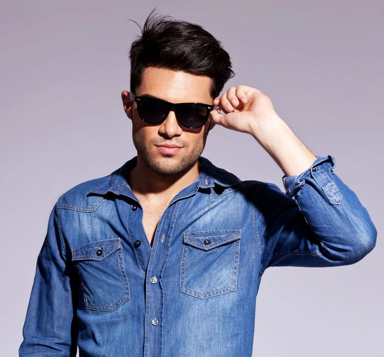 Uomo con camicia di jeans blu che si sistema gli occhiali da sole neri su uno sfondo viola chiaro