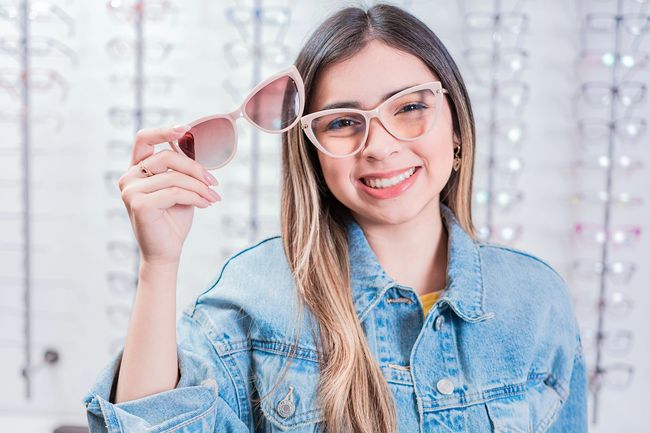Persona sorridente che tiene accanto al viso degli occhiali da sole rosa, indossa una giacca di jeans e occhiali trasparenti.