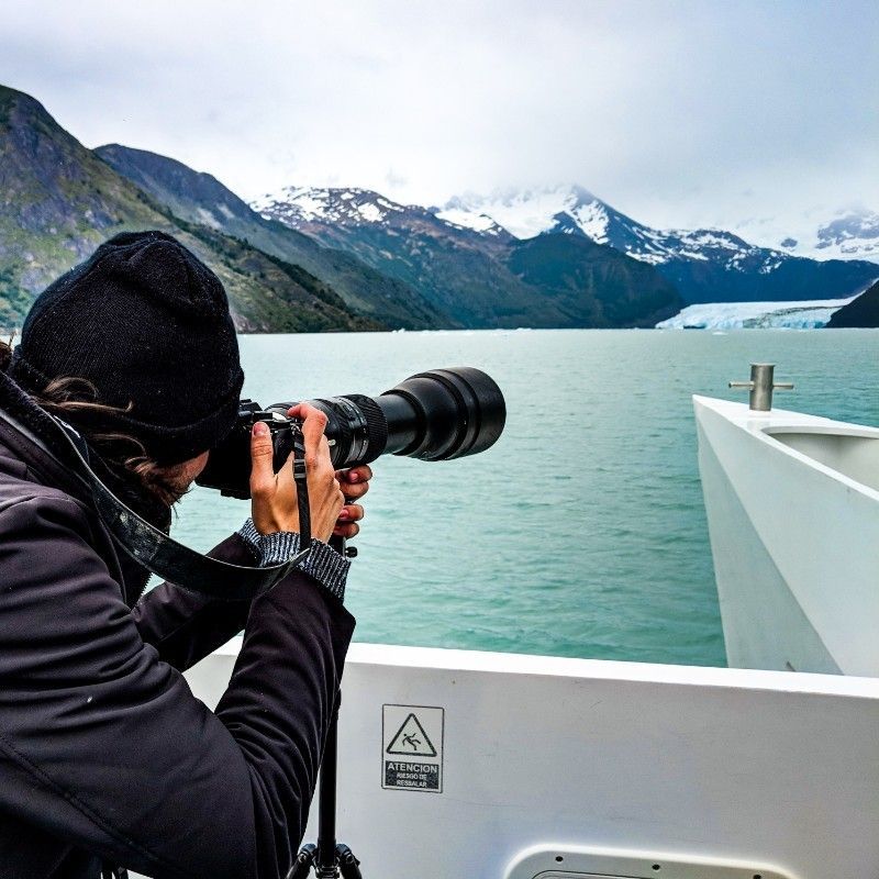 Photographer on boat taking photo of mountains and glacier, overcast sky.