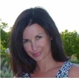Woman with long, dark hair smiles, wearing a patterned top, outdoors with greenery in the background.