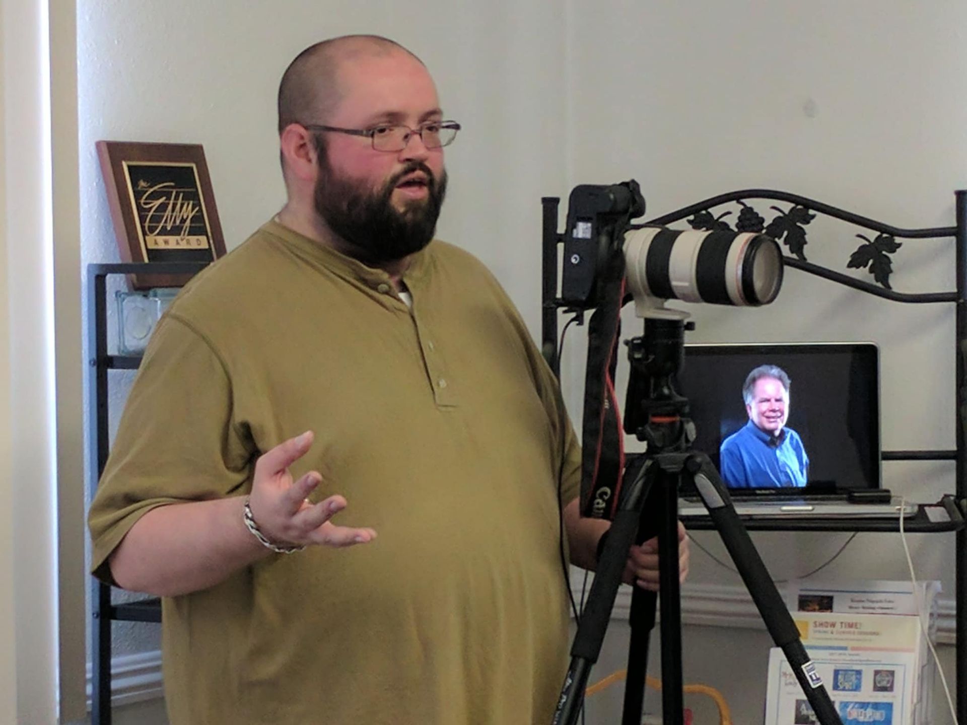 A man with a beard and glasses gestures near a camera mounted on a tripod, with a laptop showing a portrait in the back.