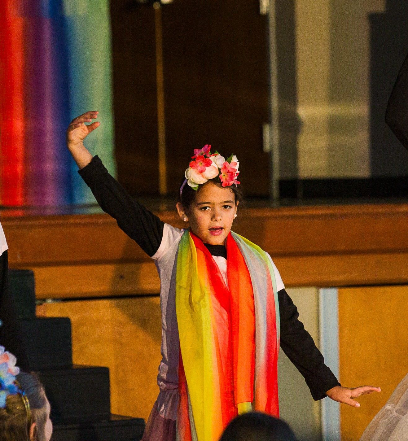Child in colorful costume and flower crown with one arm raised on a stage