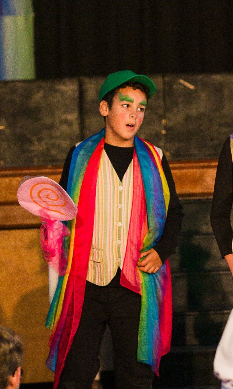 Boy in a green cap and rainbow scarf holding a large pink lollipop prop on stage, wearing a cream vest.