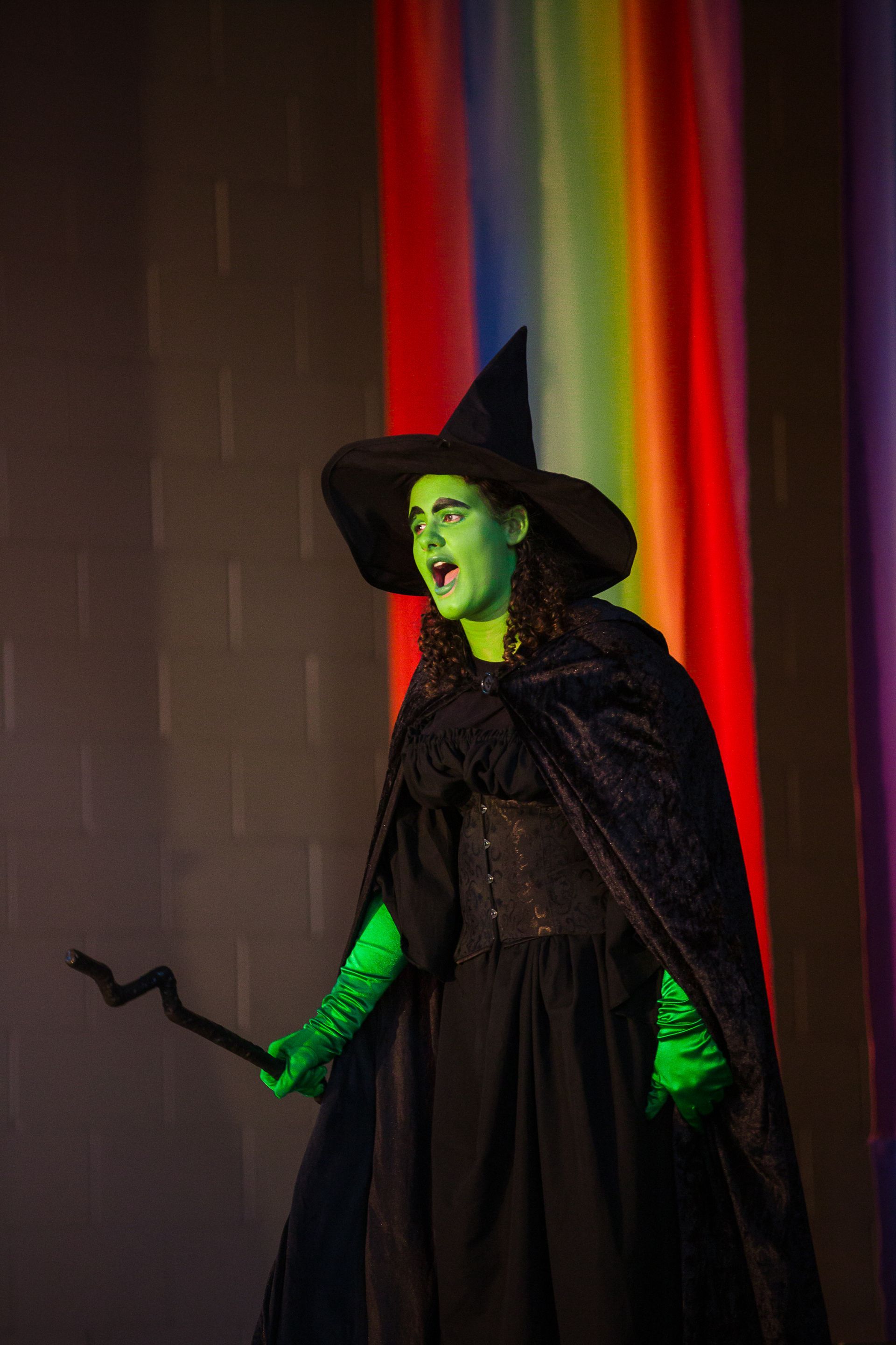 Costumed green-faced witch in black hat and cloak, holding a wand against a rainbow backdrop