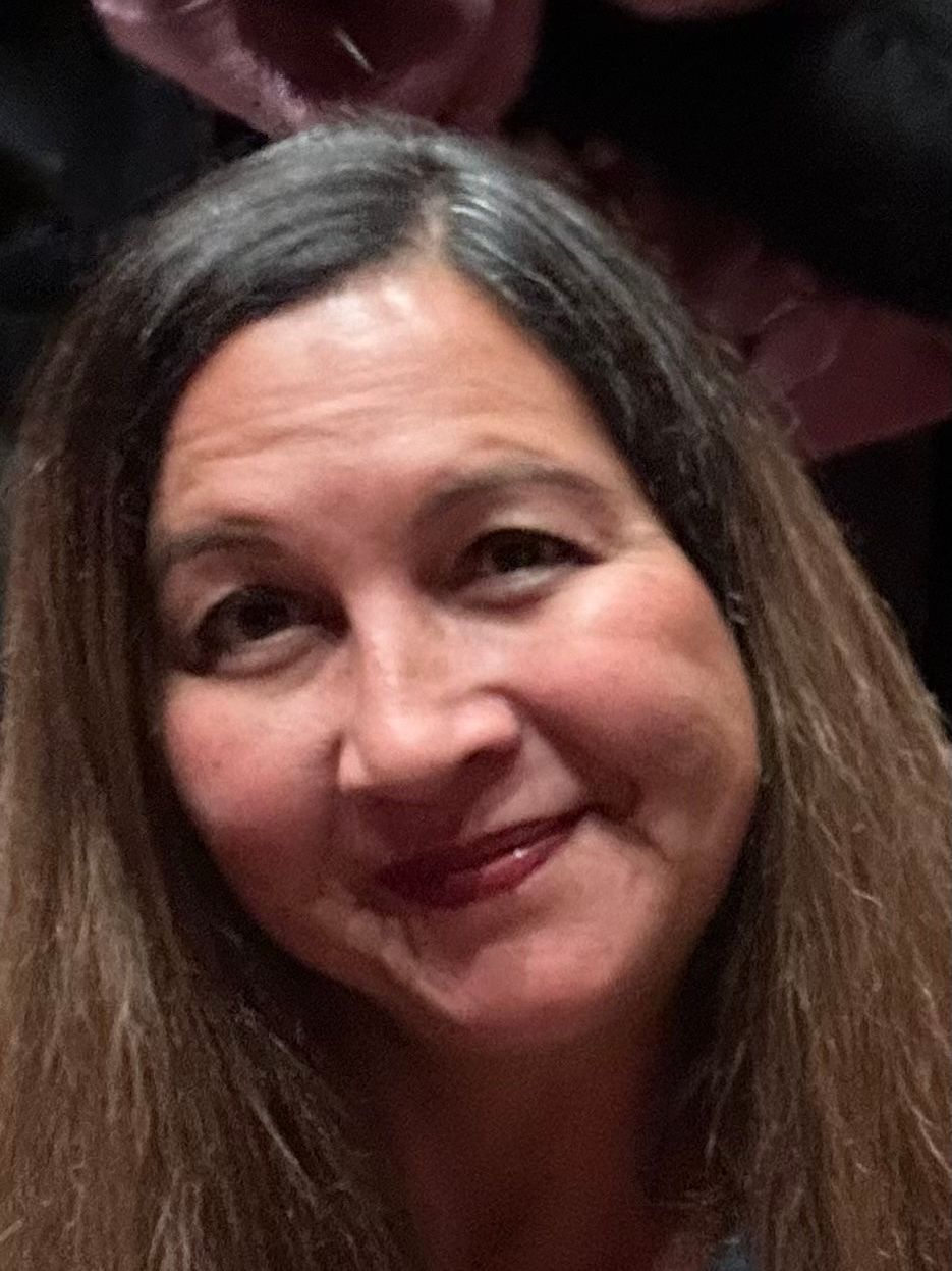 A smiling person with long, dark, wavy hair looks toward the camera.