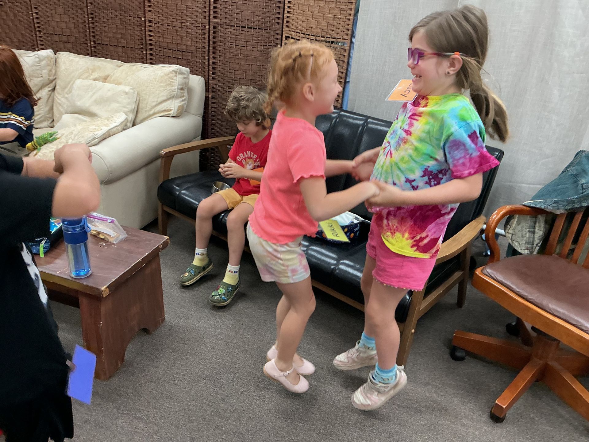 Two children stand indoors, holding hands, while another child sits on a sofa in the background.