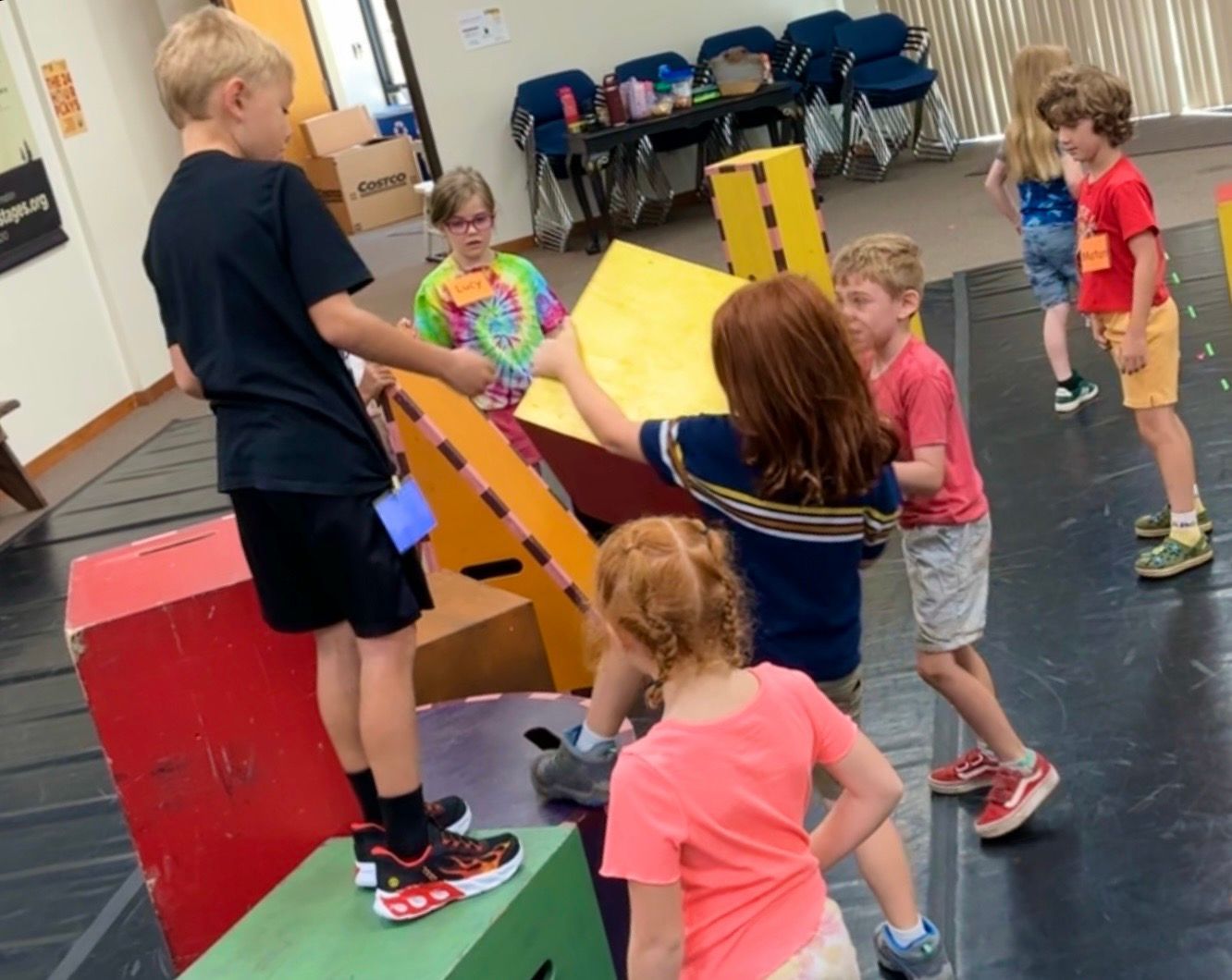 Children in a gym stacking colorful foam blocks while one child stands on top looking down