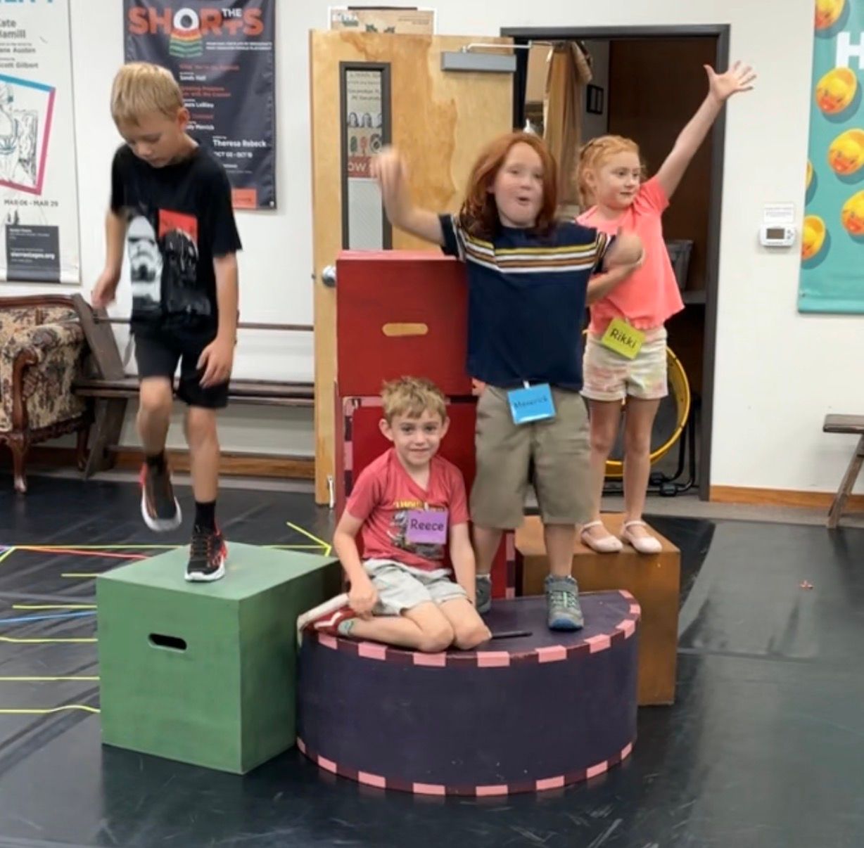 Children posing on colorful boxes and platforms in a classroom, one with arms raised in celebration