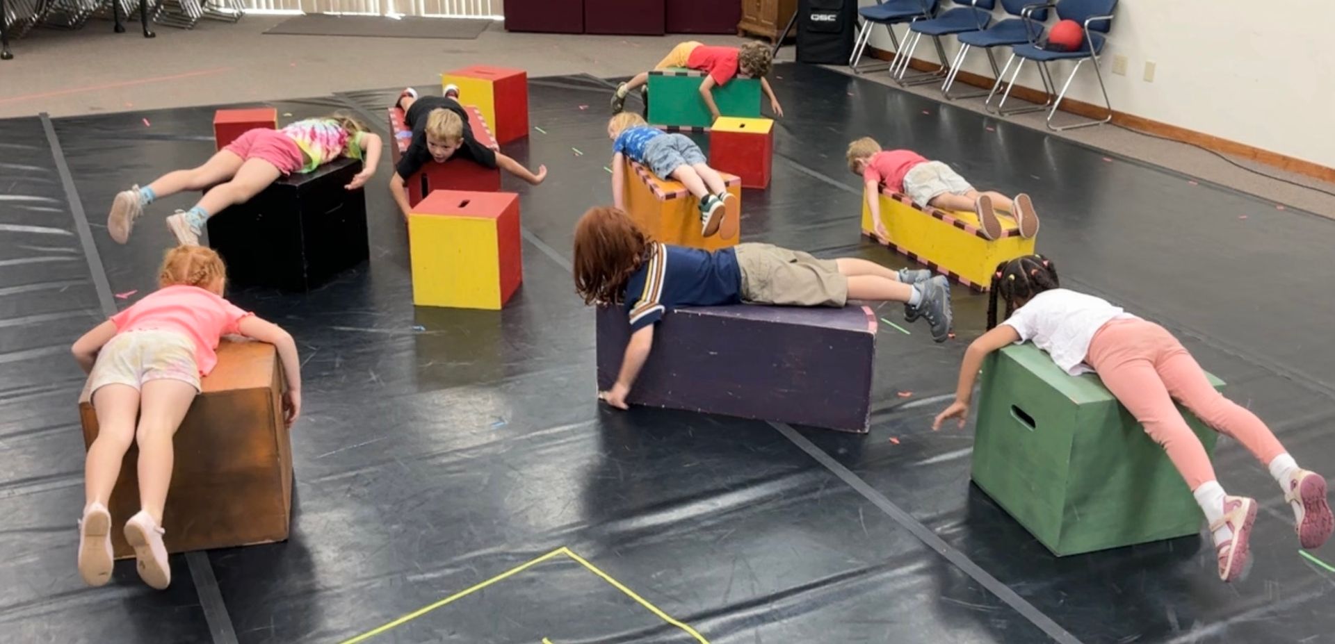 Children lying over colorful foam blocks in a gym, practicing a group balance or stretching exercise