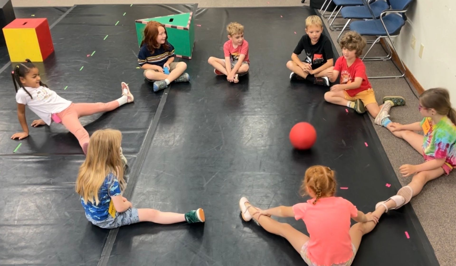 Children sitting in a circle on gym mats, with a red ball in the center.