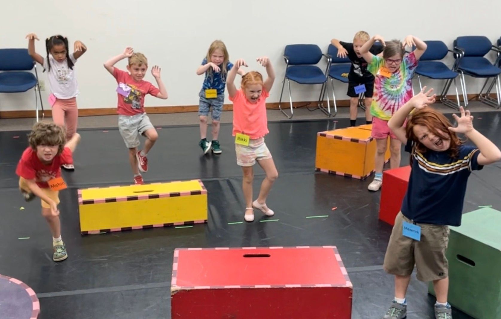 Children doing a dance or exercise routine in a room with colorful boxes and chairs along the wall
