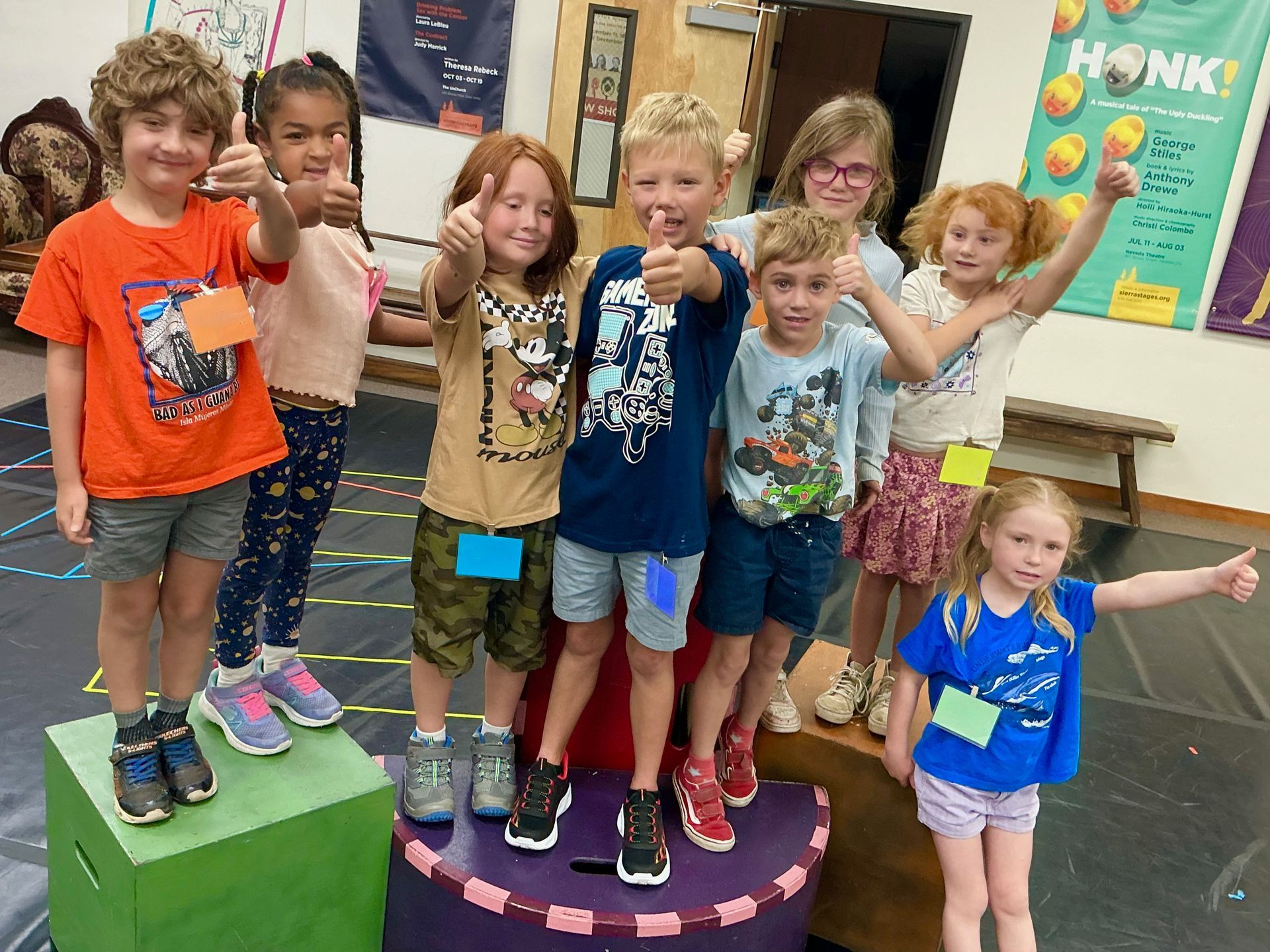 Seven children posing on colorful podiums in a classroom, smiling and waving.