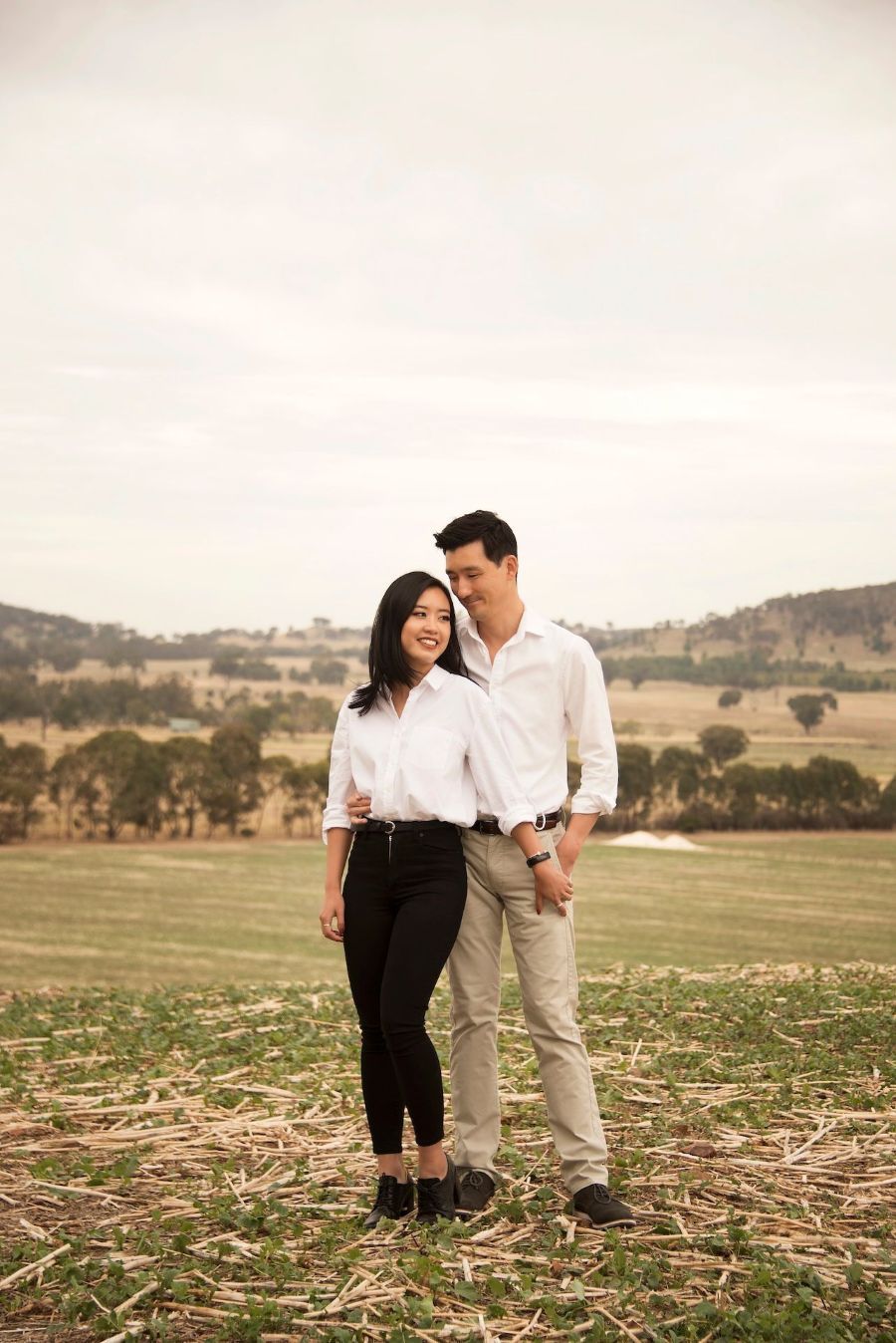 Couple in white shirts embraces in a field, looking toward each other. Hills and sky in the background — Dale Wright Portrait Maker in Shepparton, VIC