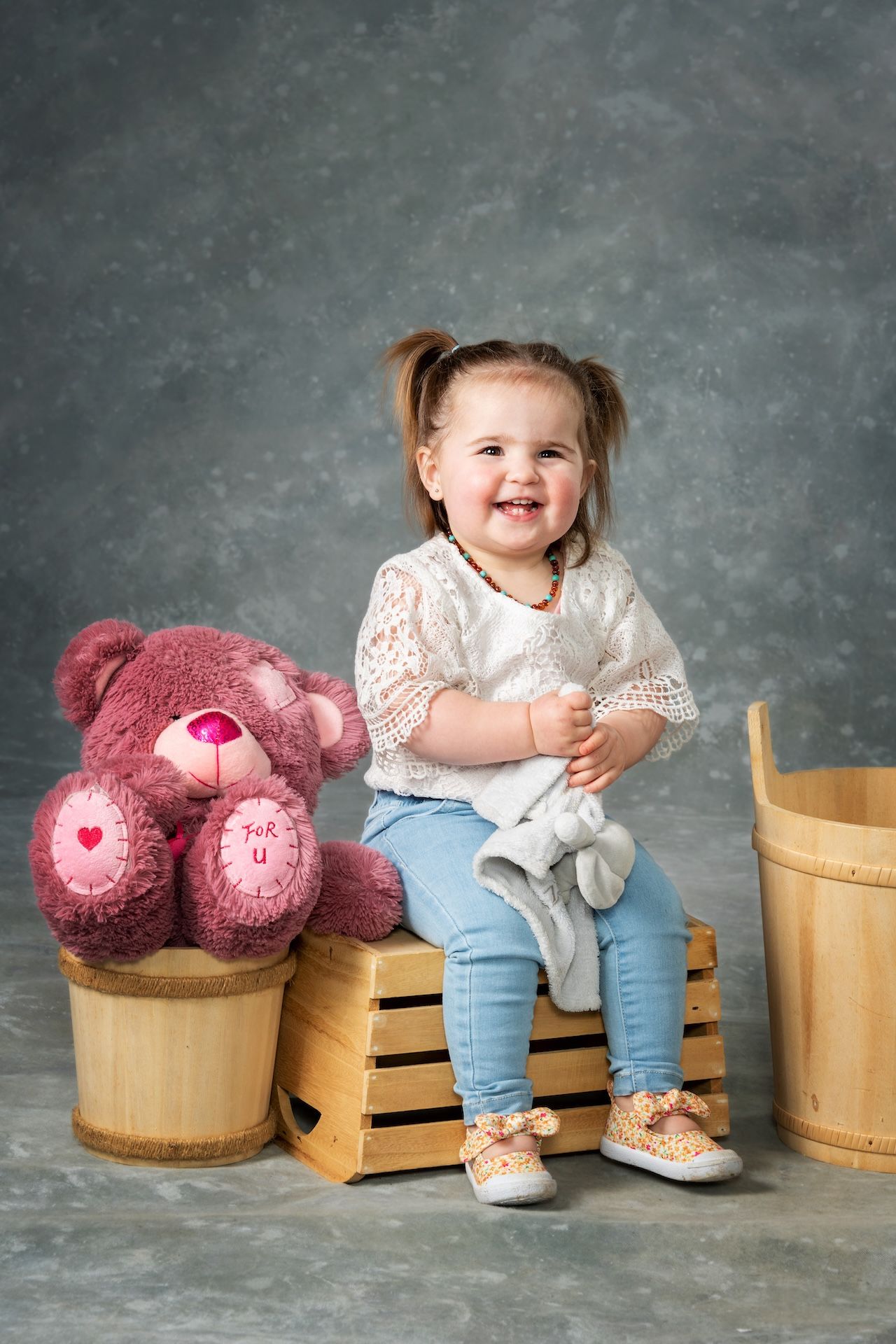 Smiling toddler with pigtails sits on a crate, holding a plush toy. A large teddy bear and wooden bucket are nearby — Dale Wright Portrait Maker in Wangaratta, VIC