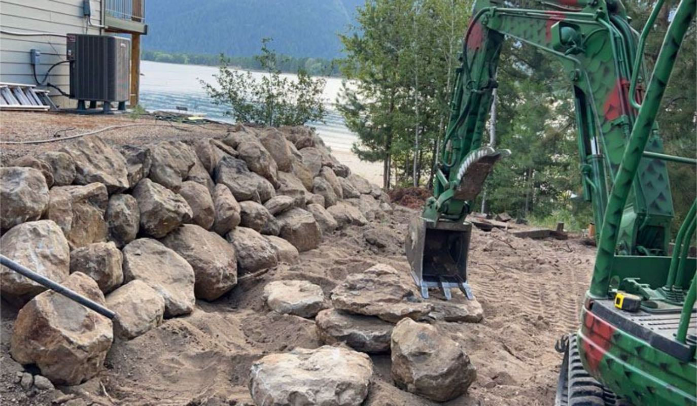 Green excavator building a stone retaining wall near water.