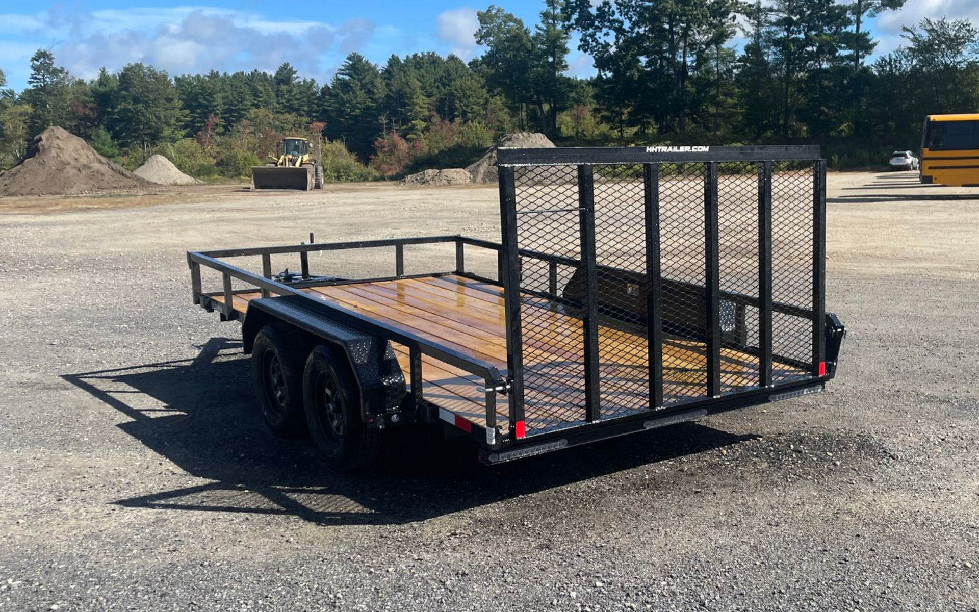 Black utility trailer with mesh sides on a gravel lot; trees in background.