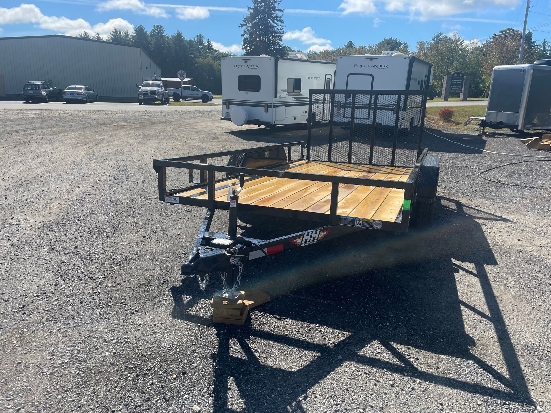 Black utility trailer with wooden deck on gravel lot, trailers and buildings in background.