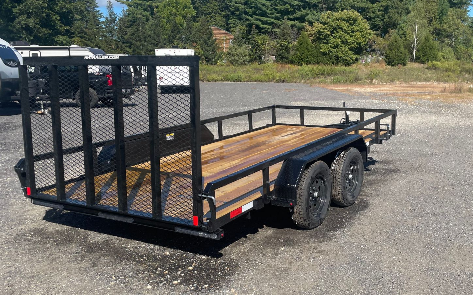Black utility trailer with wooden deck, black mesh sides, and tandem wheels parked on gravel.
