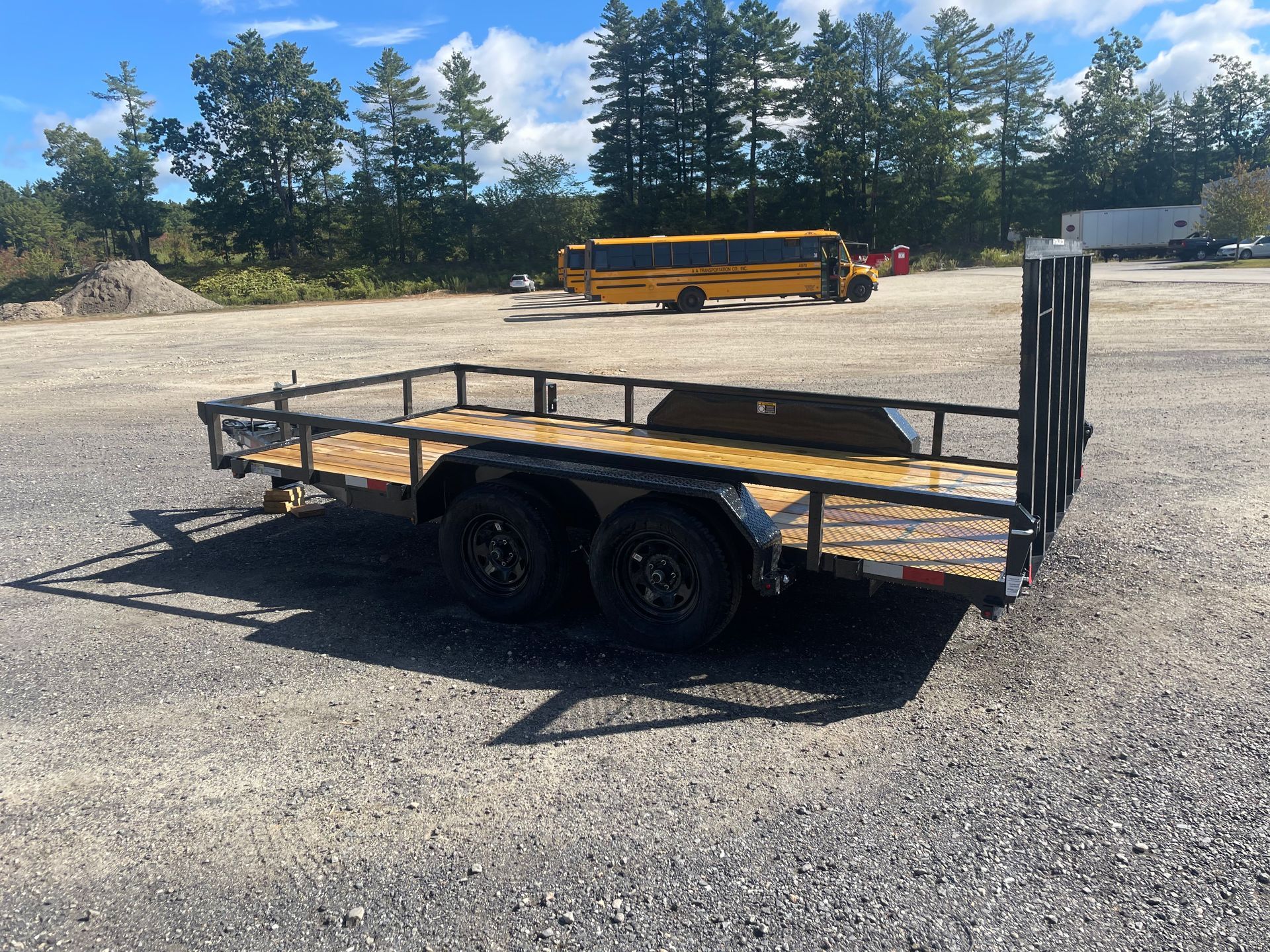 A black flatbed trailer with a wooden deck parked on a gravel lot; a yellow school bus is visible in the background.