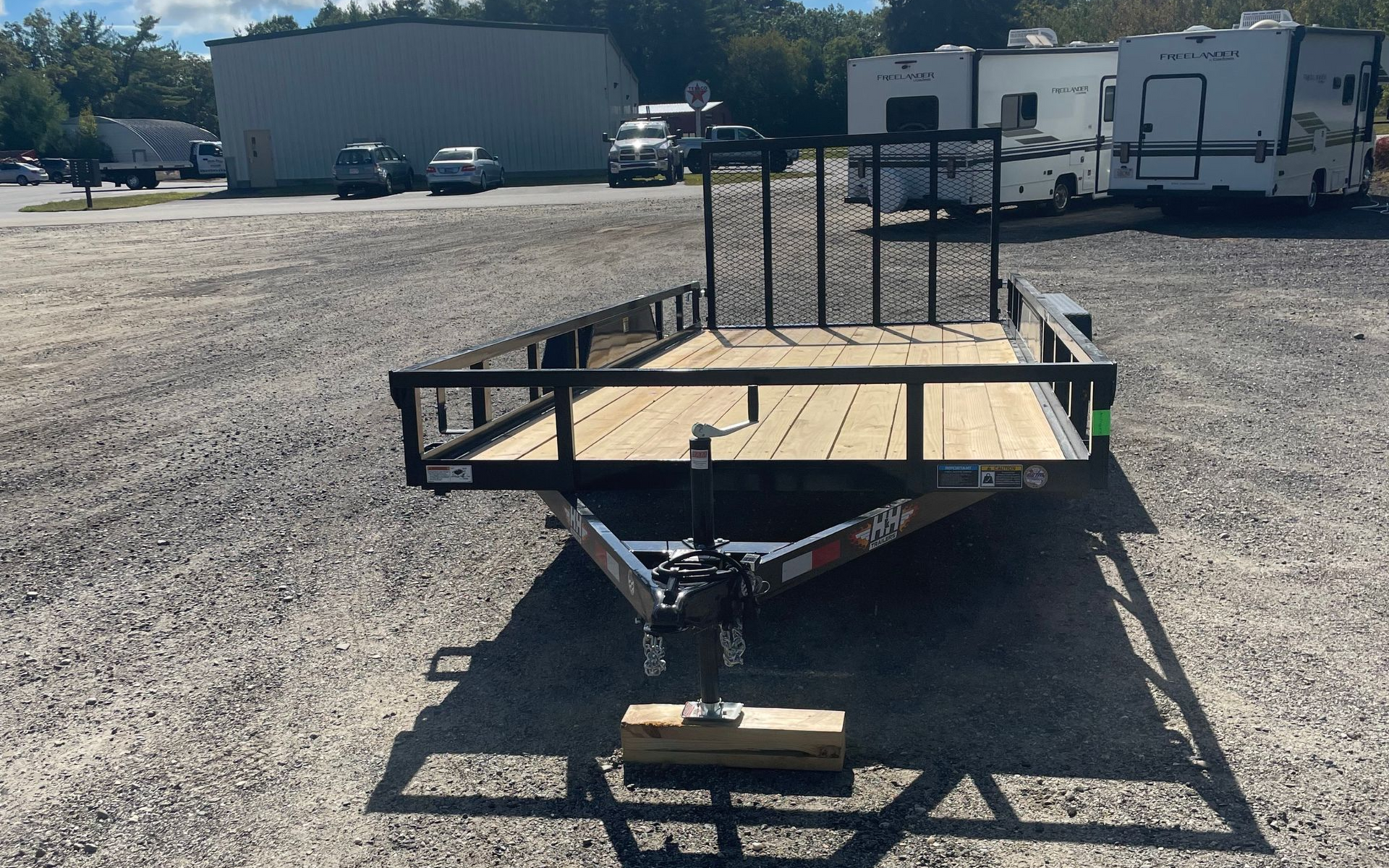 Black utility trailer with wooden deck on a gravel lot; buildings and RVs in the background.
