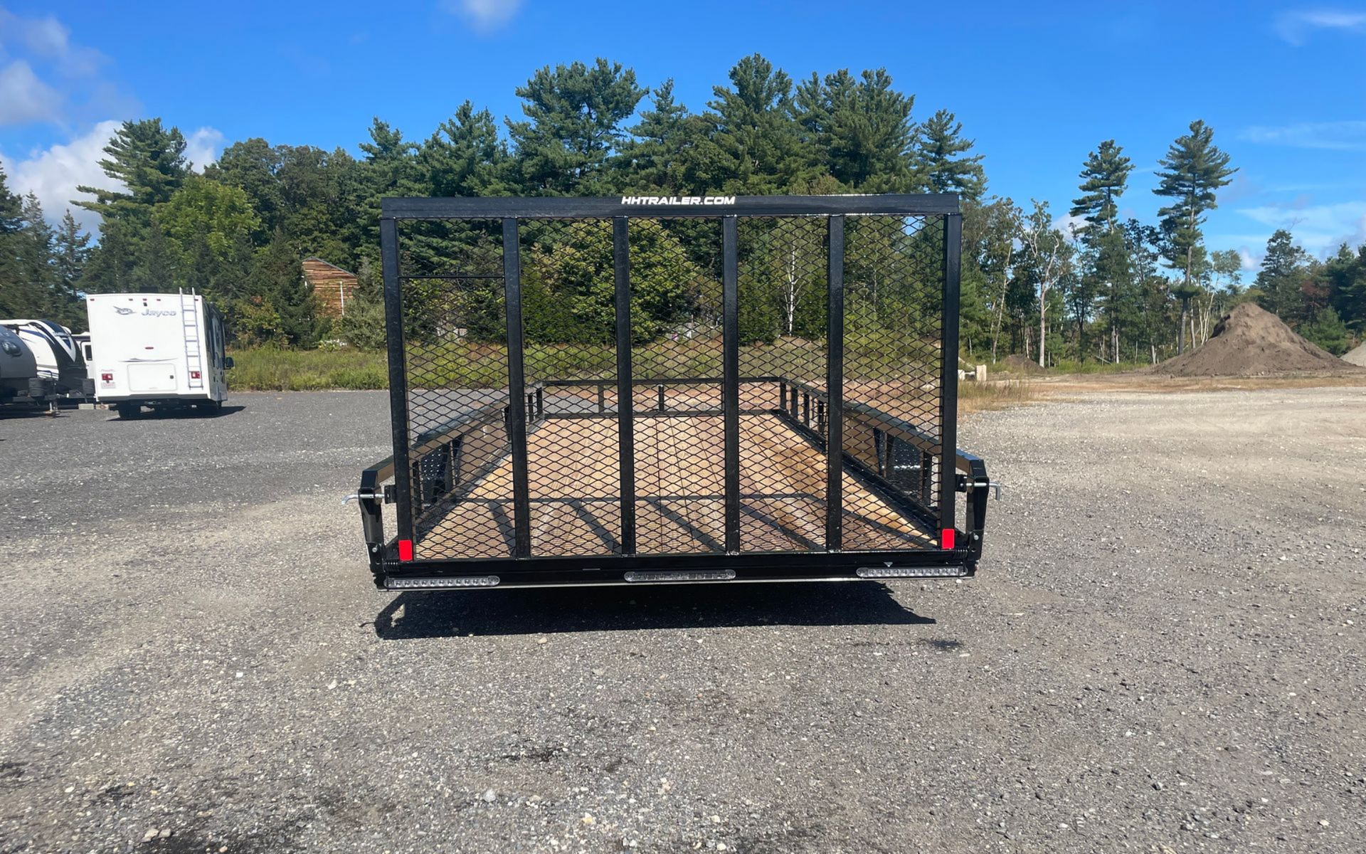 Black utility trailer with mesh sides on gravel lot under blue sky.