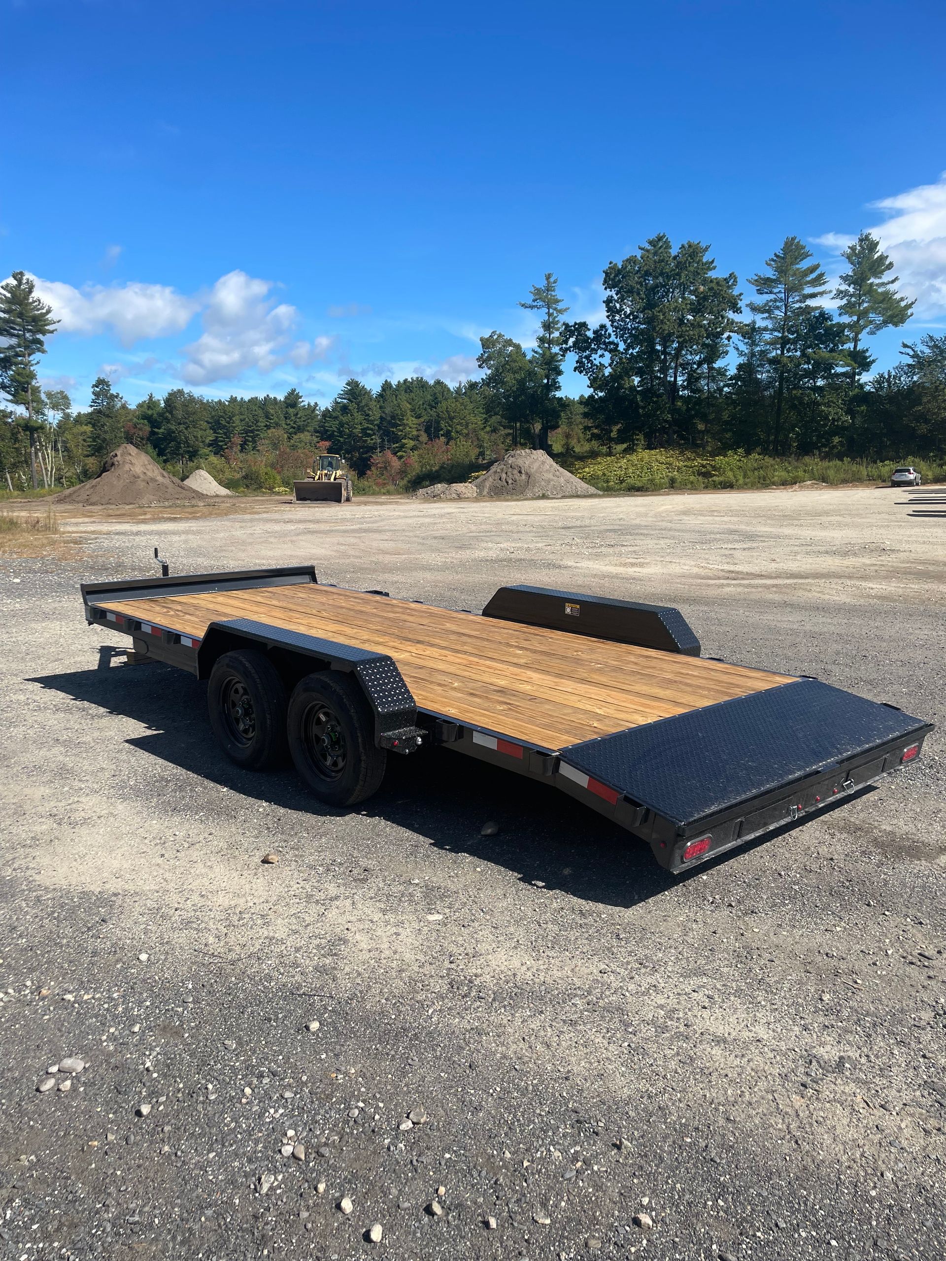 A black flatbed trailer with a wooden deck on a gravel lot under a blue sky.
