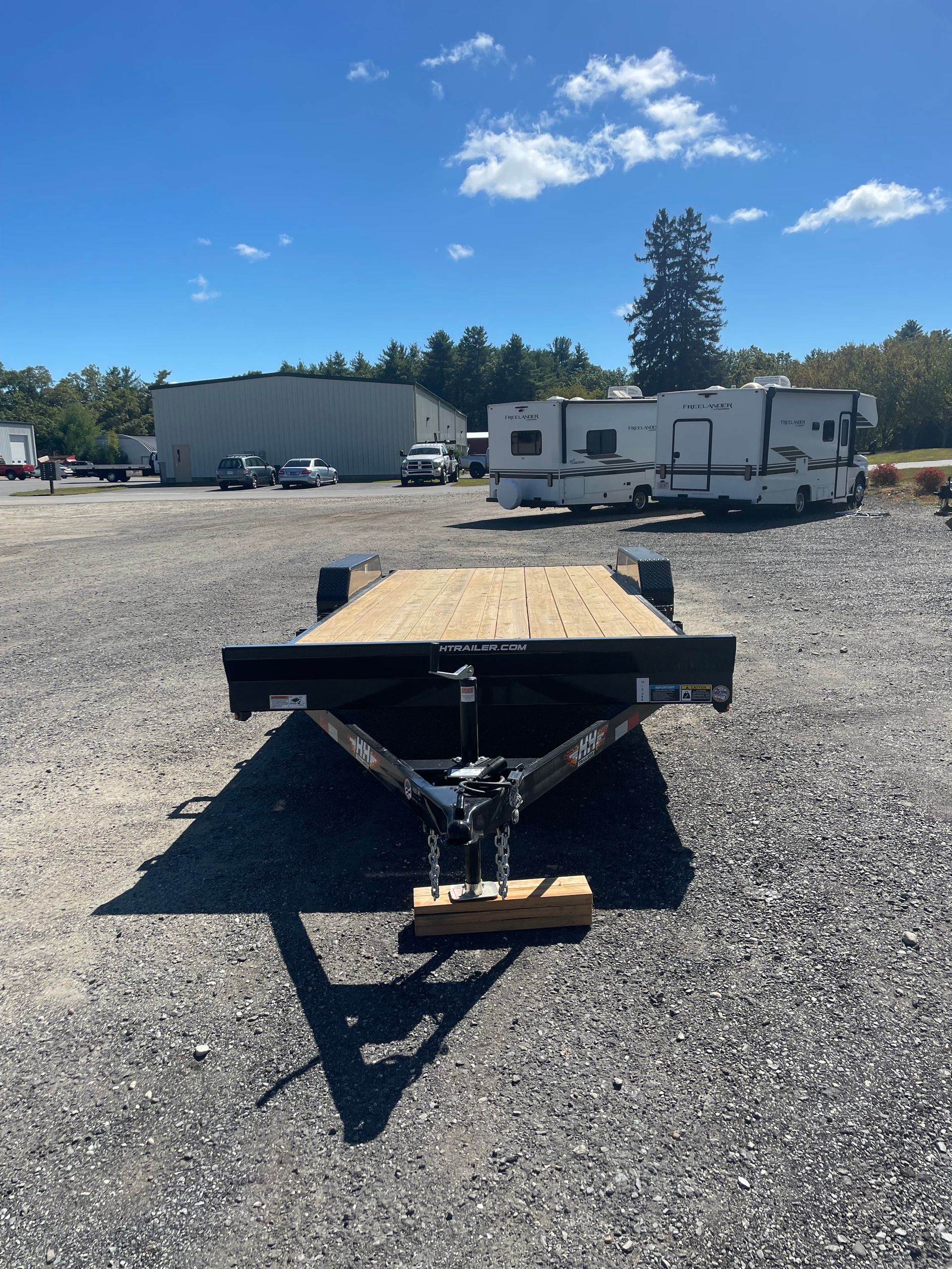 A black utility trailer with wooden deck sits on a gravel lot under a blue sky, with buildings in the background.