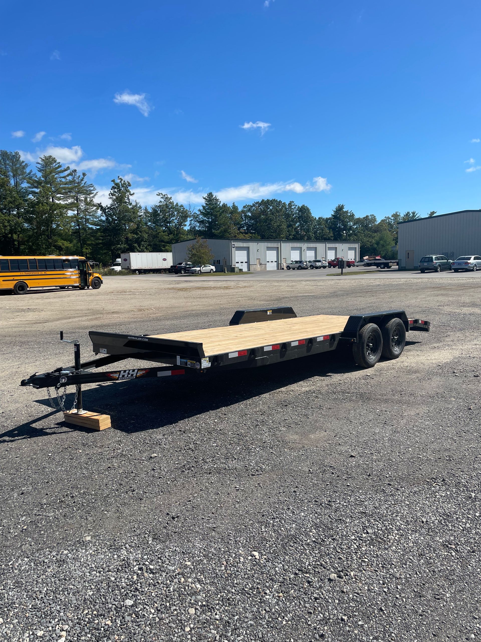A black flatbed trailer sits on gravel, under a blue sky, next to buildings.