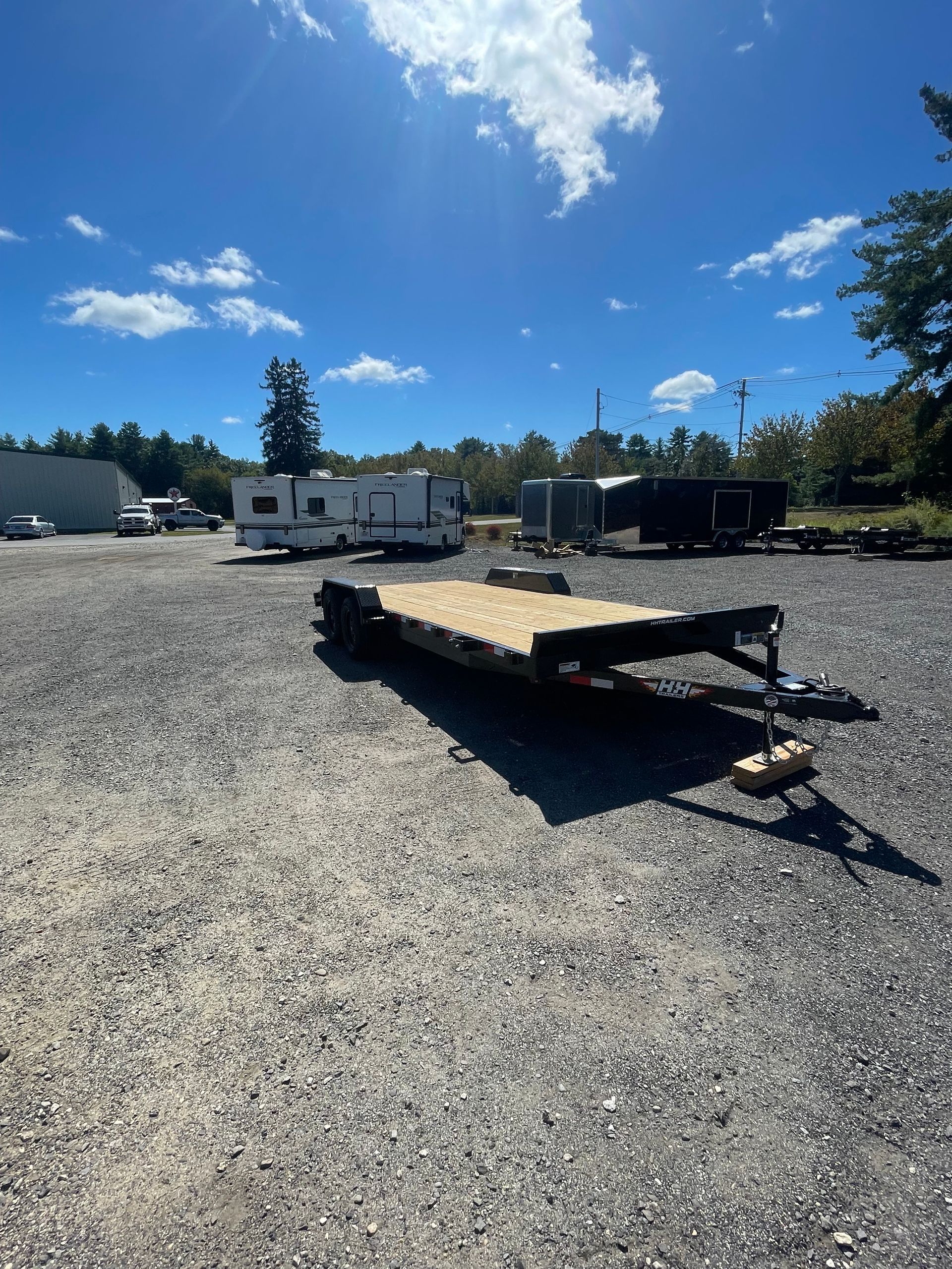 Flatbed trailer parked on gravel lot with RV and other vehicles under a blue sky.