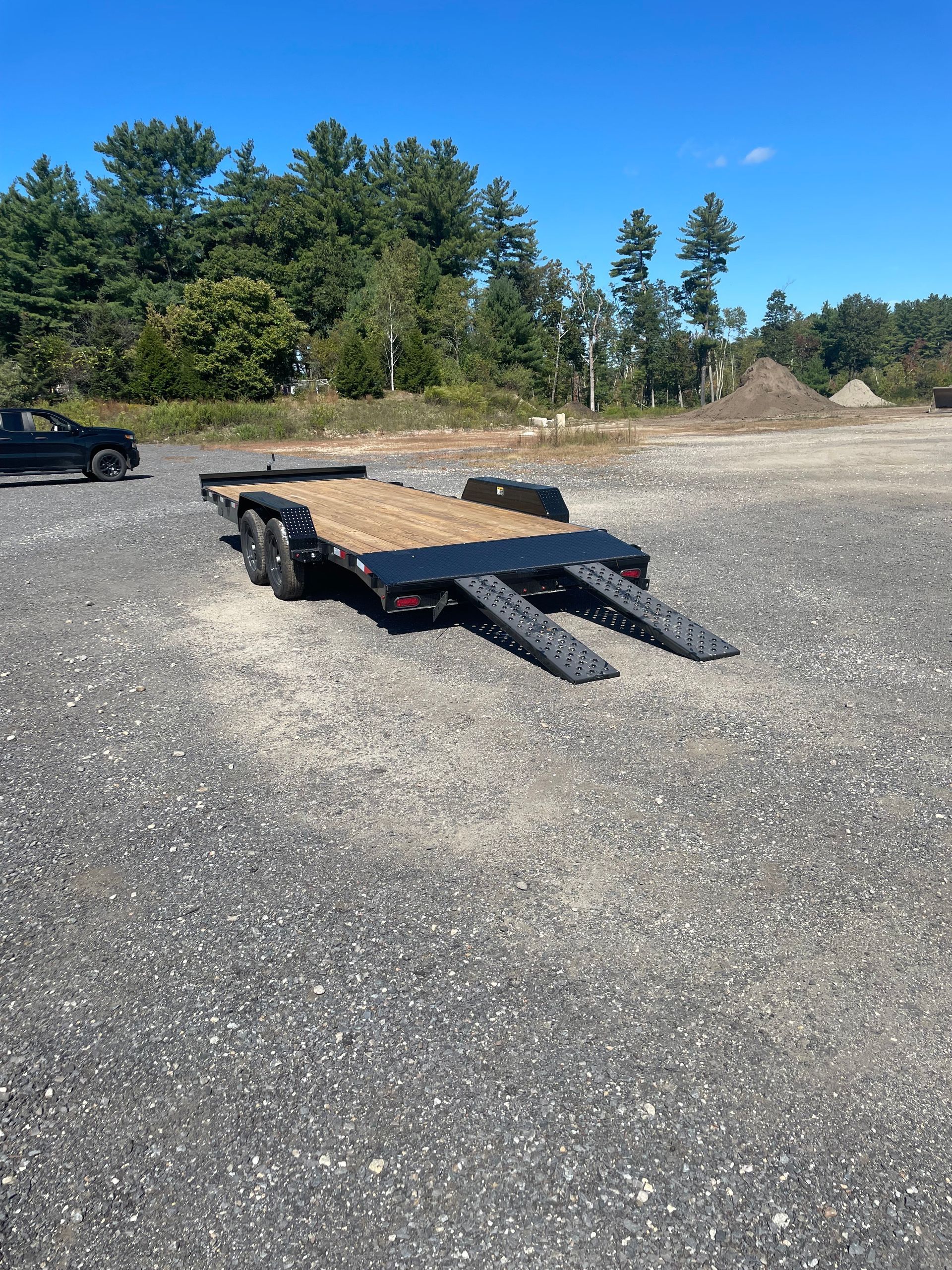 Black flatbed trailer with ramps, parked on gravel. Blue sky, trees in background.