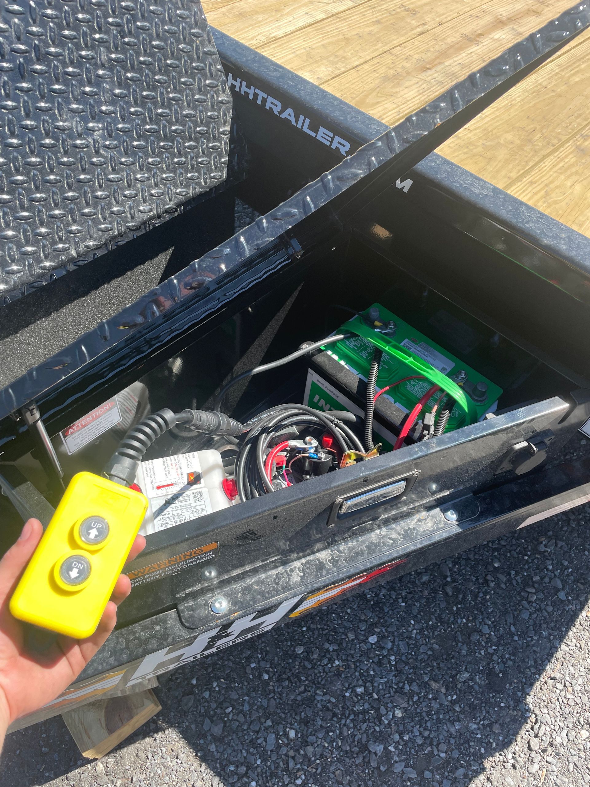 Yellow remote in hand, next to a trailer's storage compartment holding a battery and wiring.