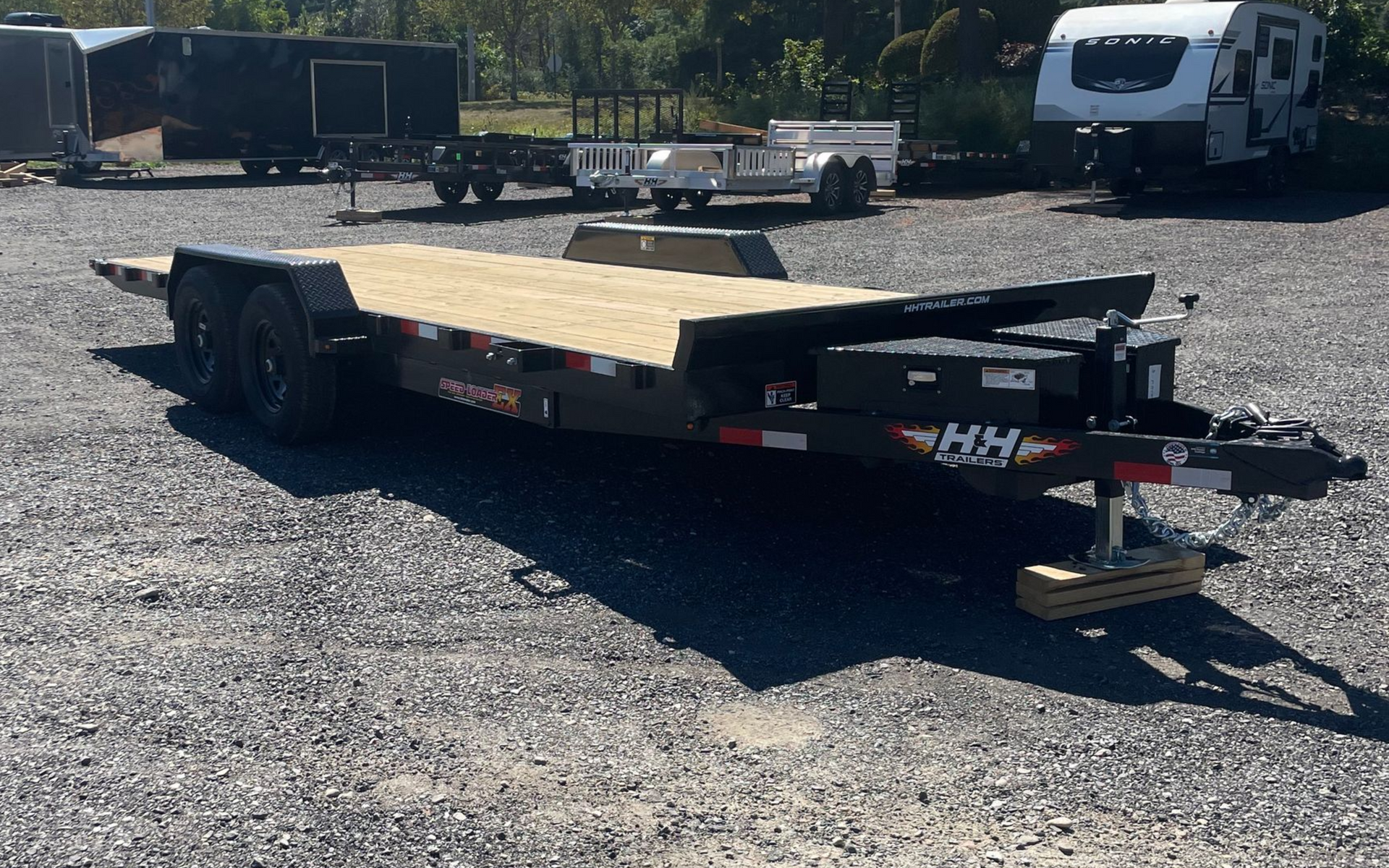 Black flatbed trailer with wooden deck on gravel. Other trailers and vehicles in background.
