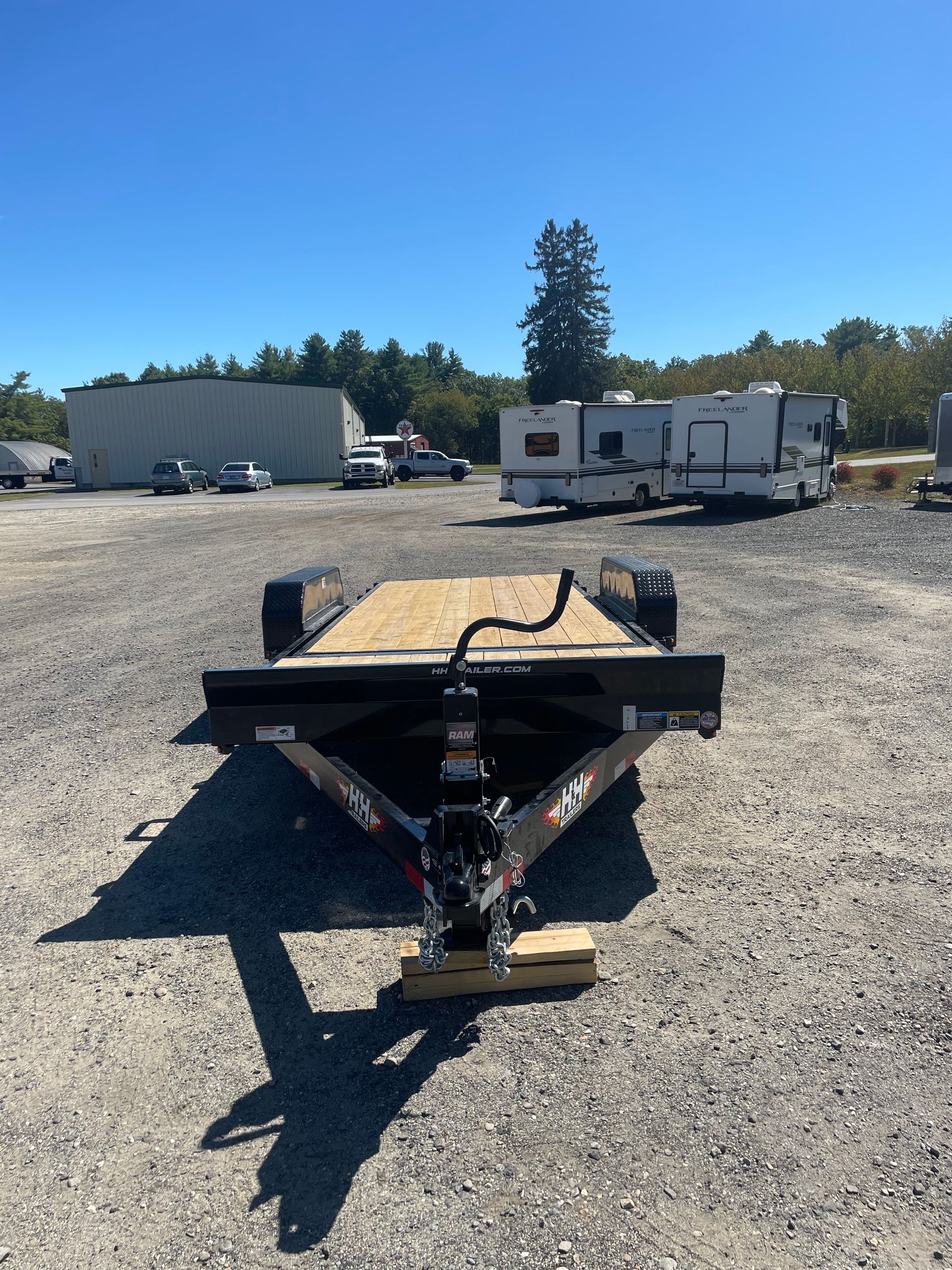 Black trailer on gravel, with wooden deck, parked outdoors under a blue sky.