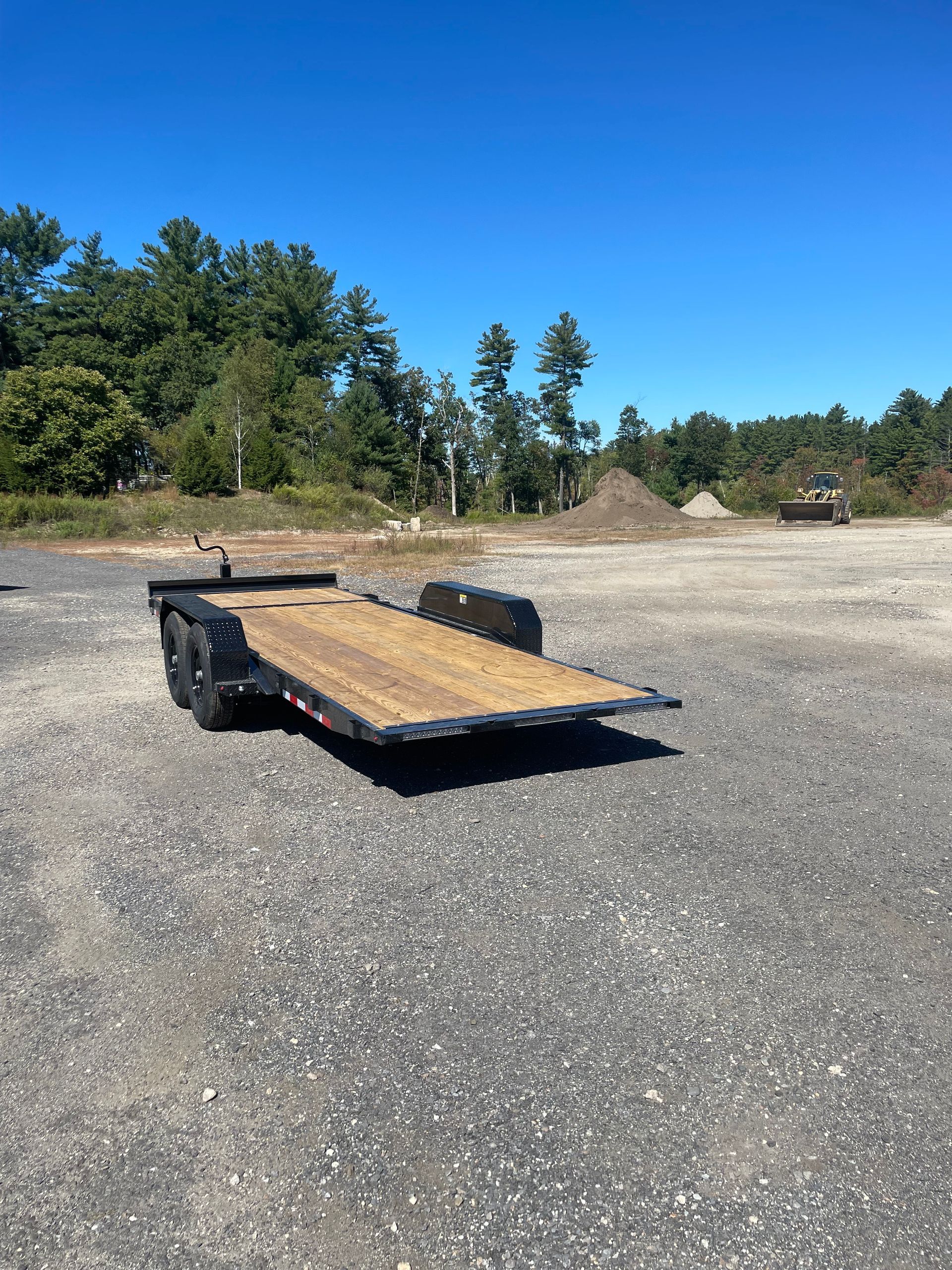 Black trailer with wooden deck parked on gravel, trees in background, blue sky.