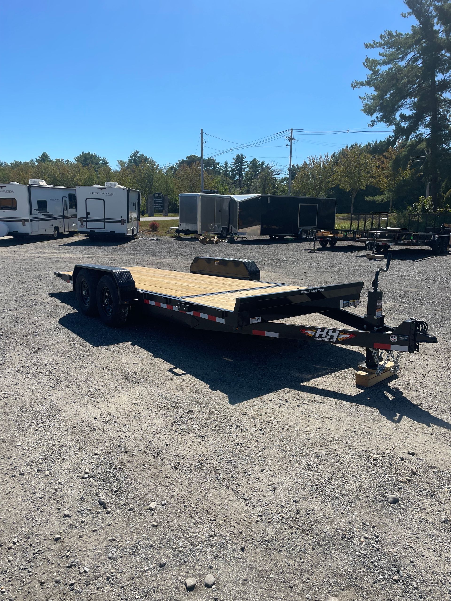 Flatbed trailer parked on gravel, with other trailers and vehicles in the background under a blue sky.