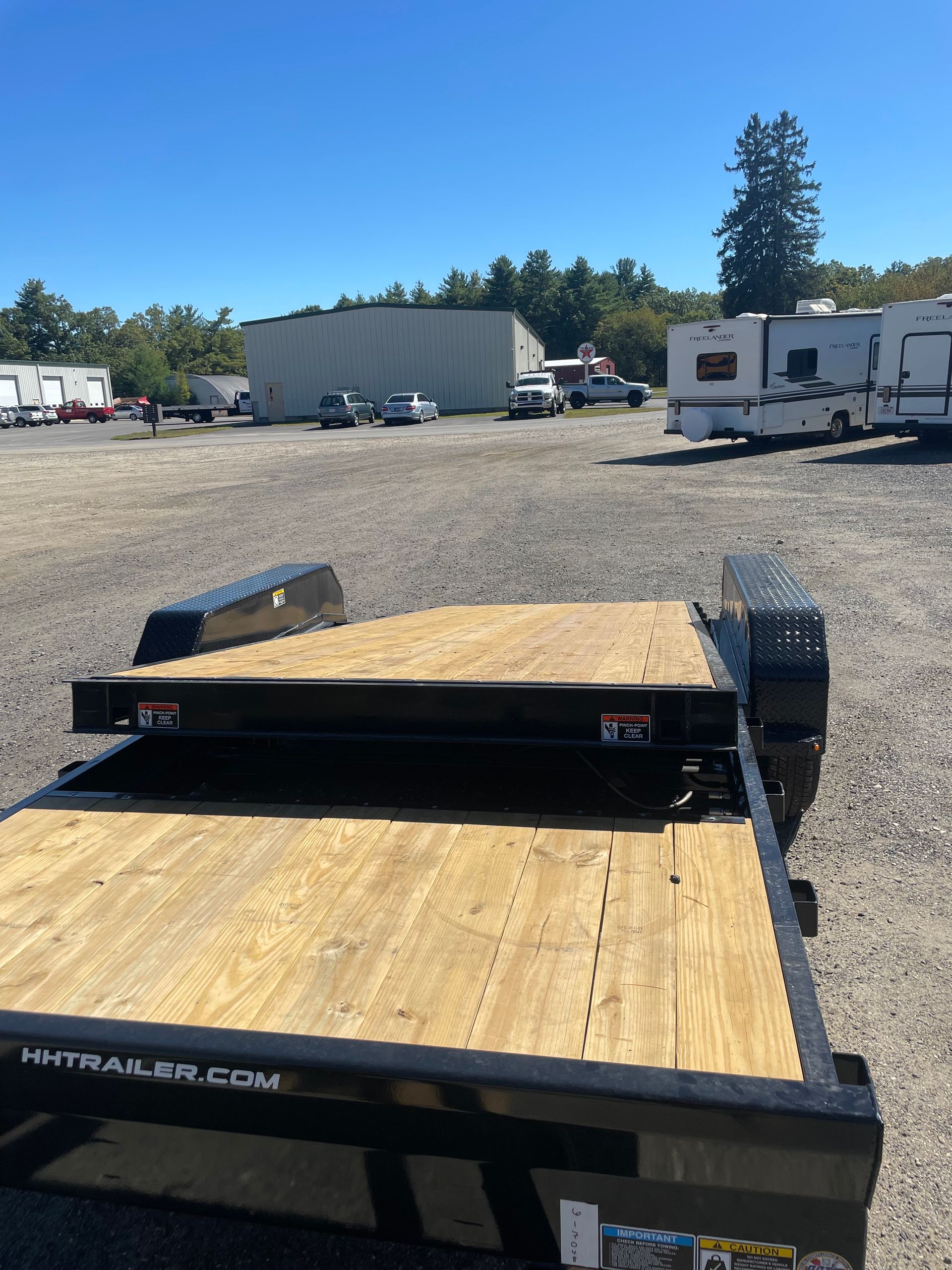 Flatbed trailer in a gravel lot with buildings and vehicles in the background. Bright blue sky.