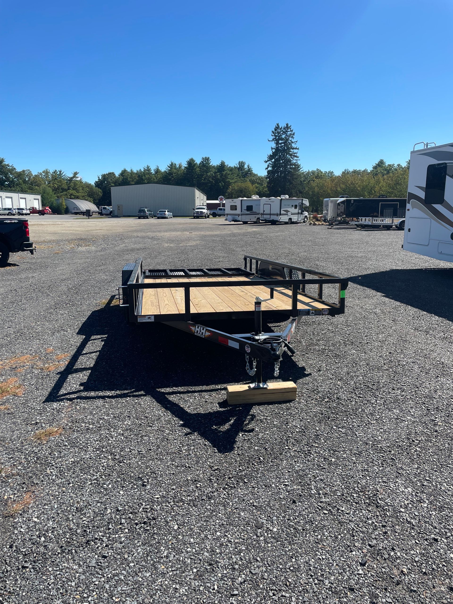 Black utility trailer on gravel lot under a blue sky, surrounded by RVs and buildings.