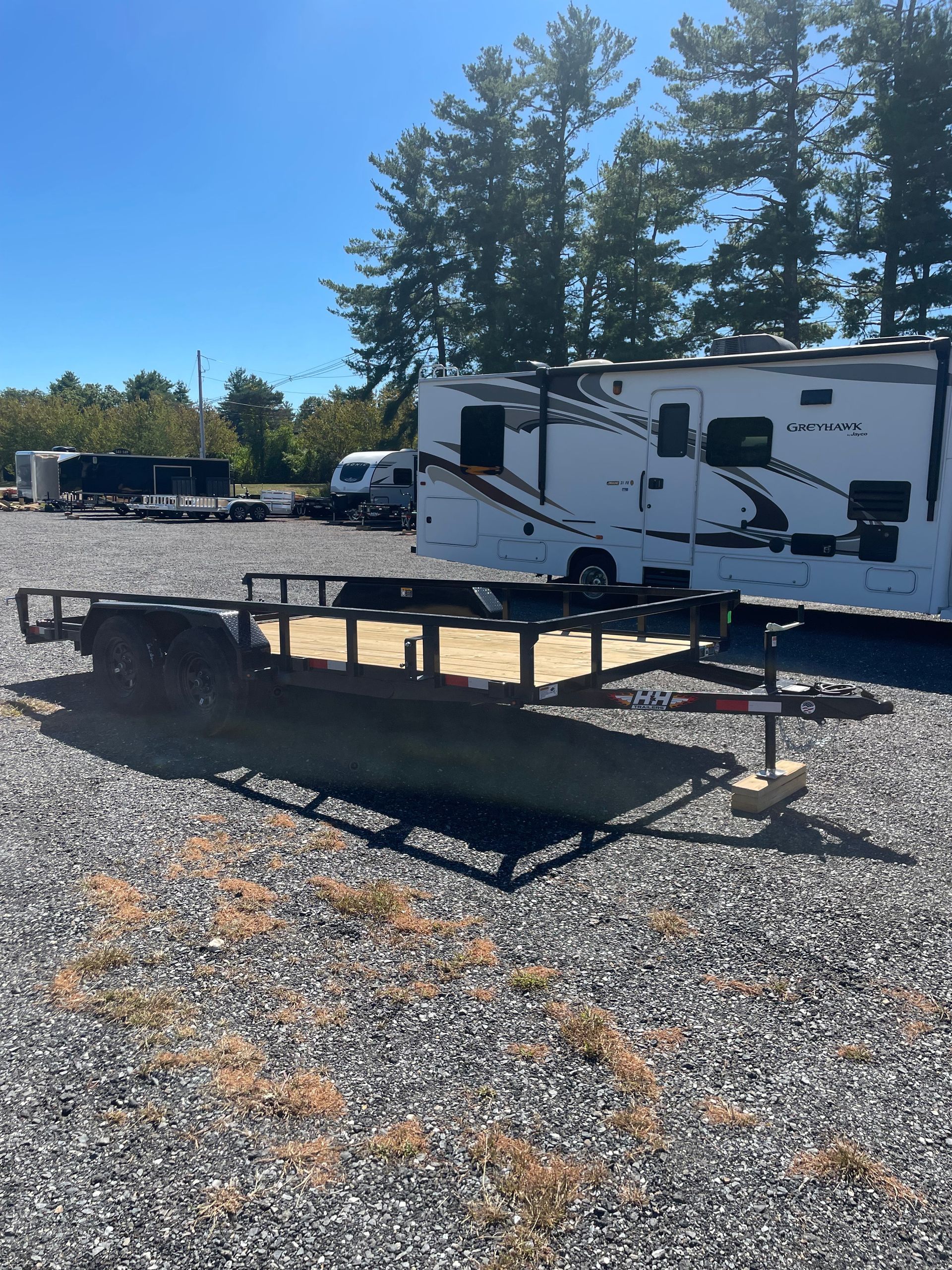 Black utility trailer on gravel, with RV and trees in the background under a blue sky.
