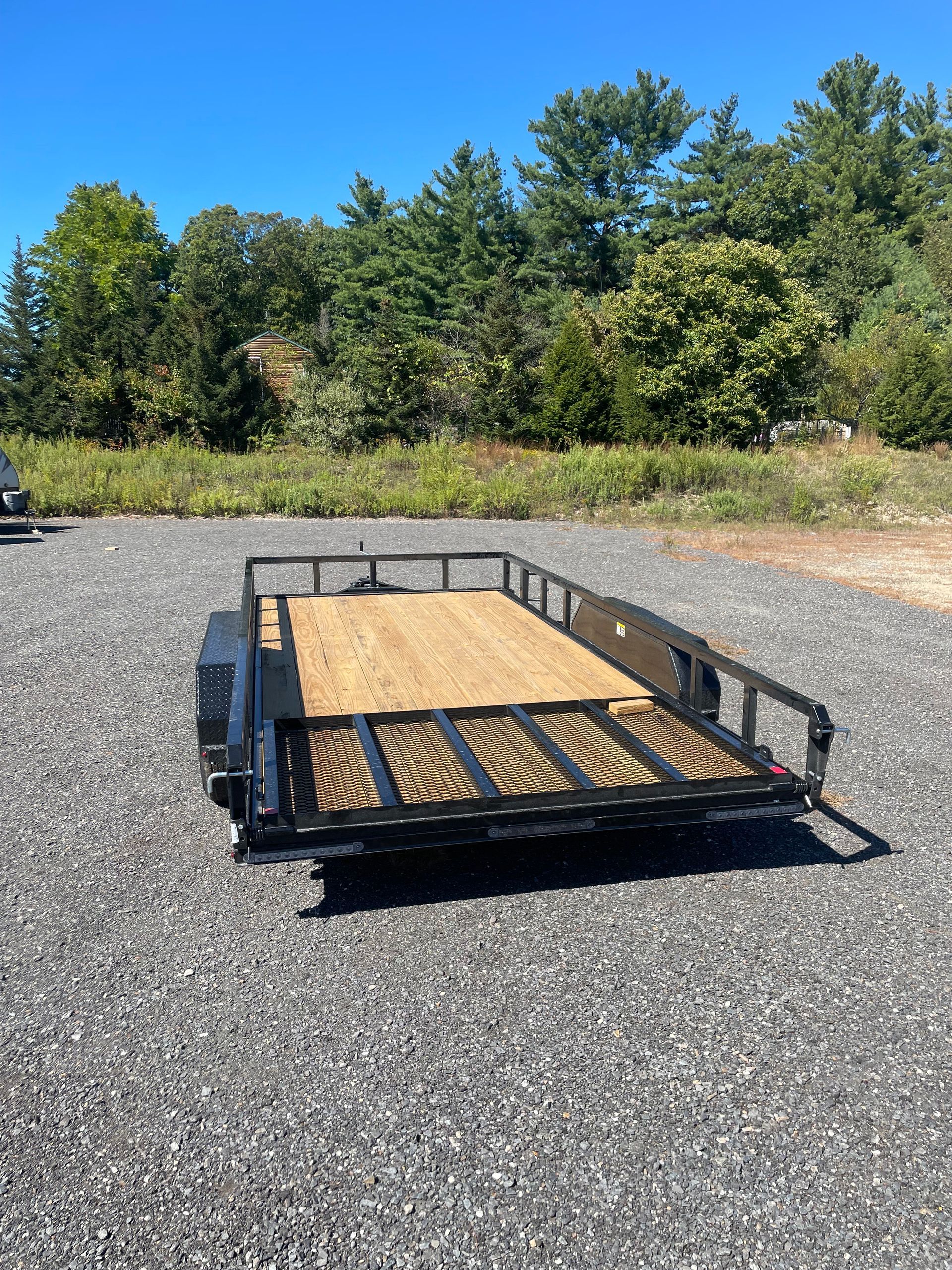 Black utility trailer with wooden floor, parked on gravel.