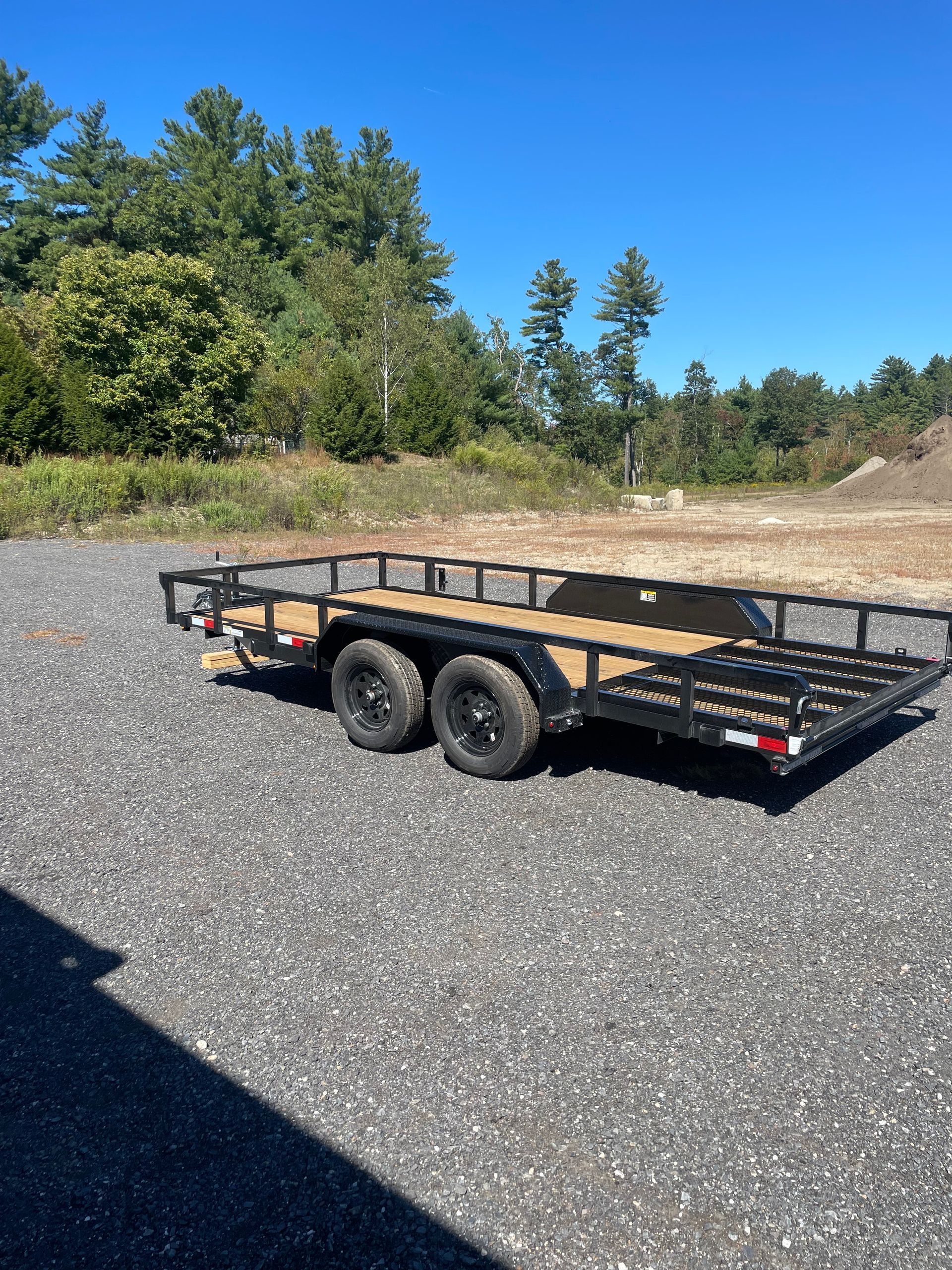 Black utility trailer with dual wheels, black metal frame, wooden deck, parked on gravel.