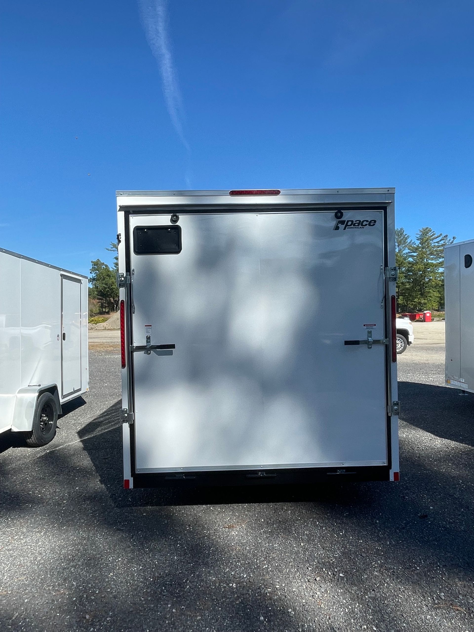 Rear view of a white enclosed cargo trailer against a blue sky, on a gravel surface, parked outside.