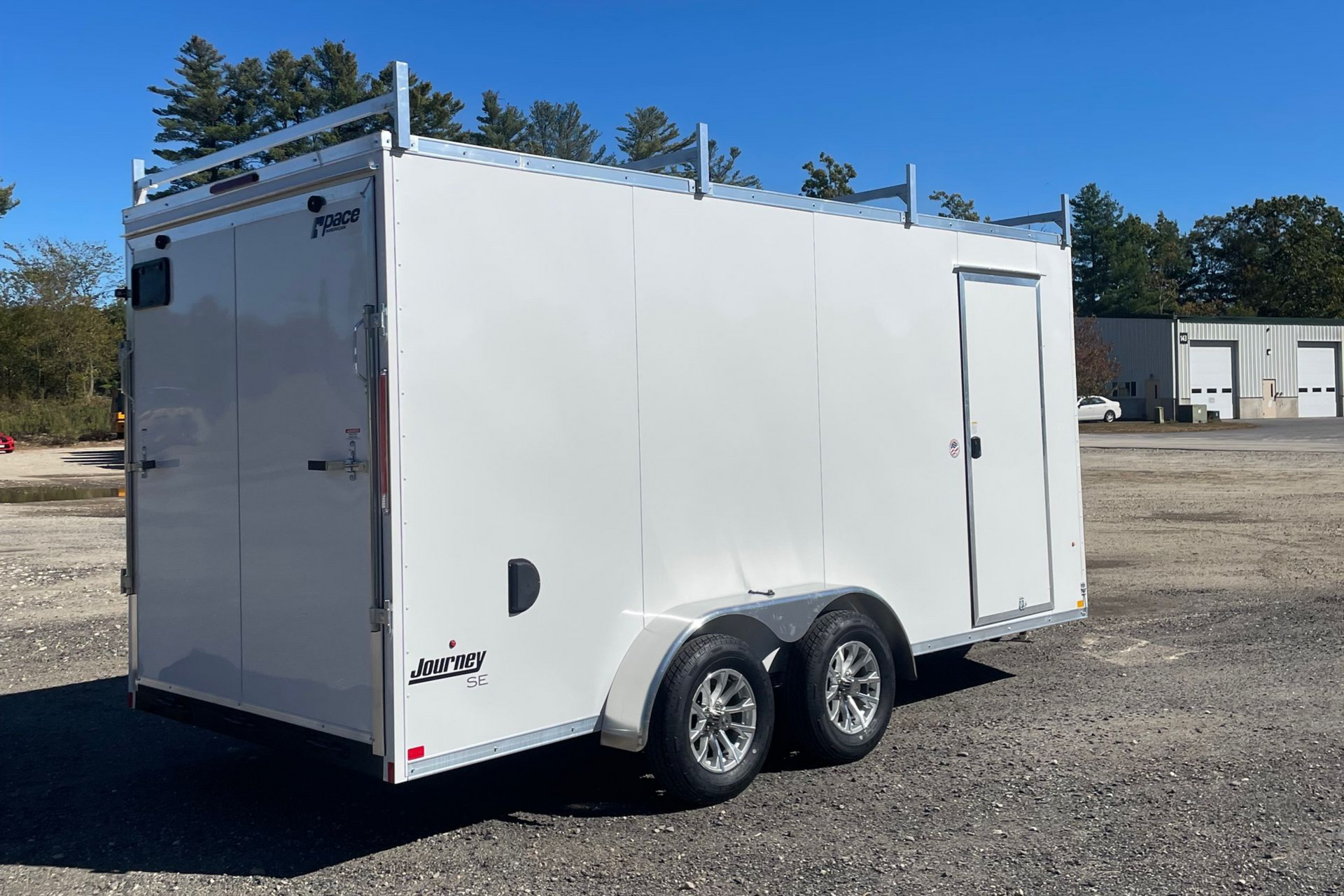 White enclosed trailer with ladder rack on the roof, side door, and dual axles, parked outdoors on a sunny day.