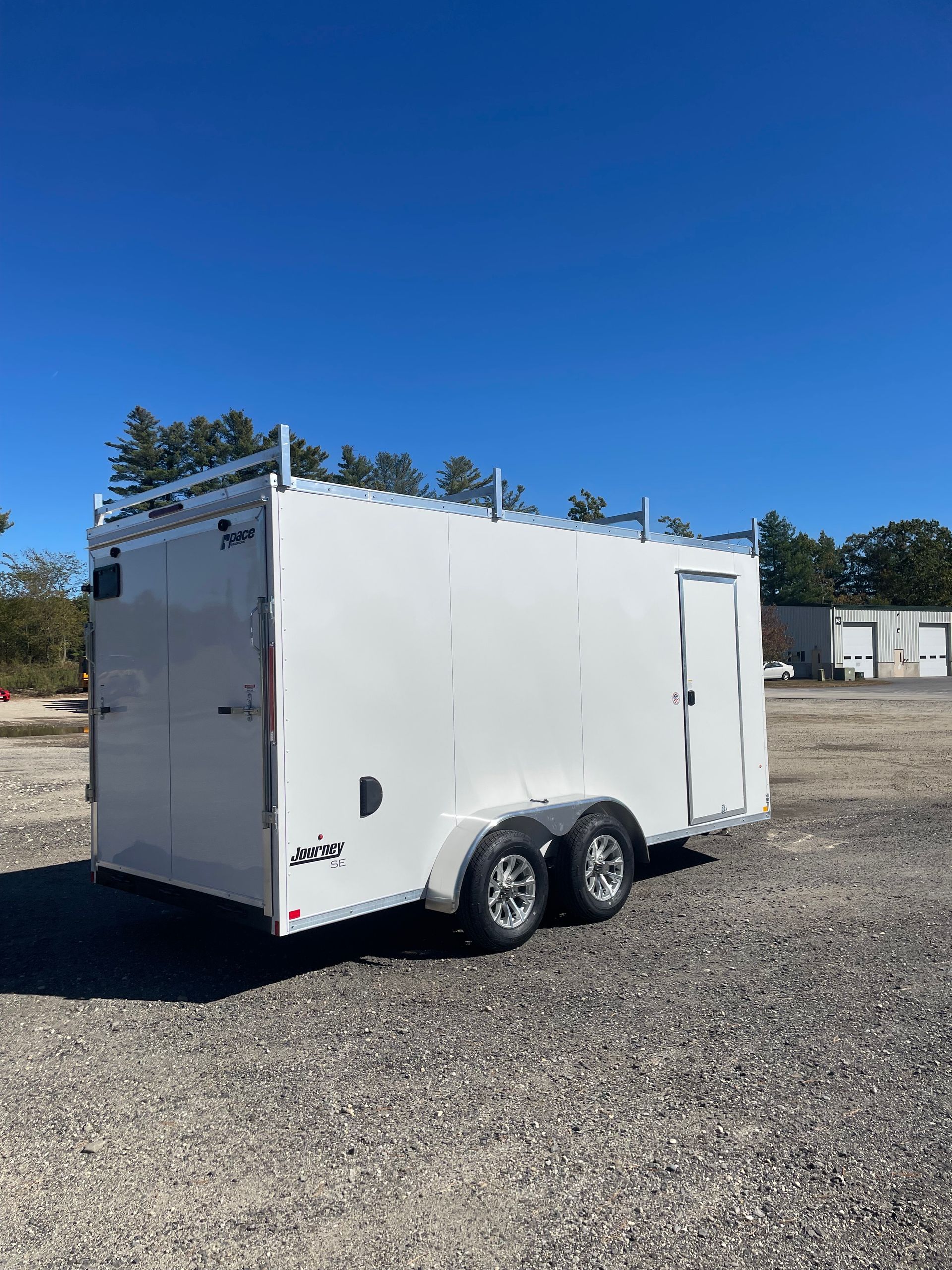 White enclosed trailer with silver trim and ladder rack, parked outdoors on gravel.
