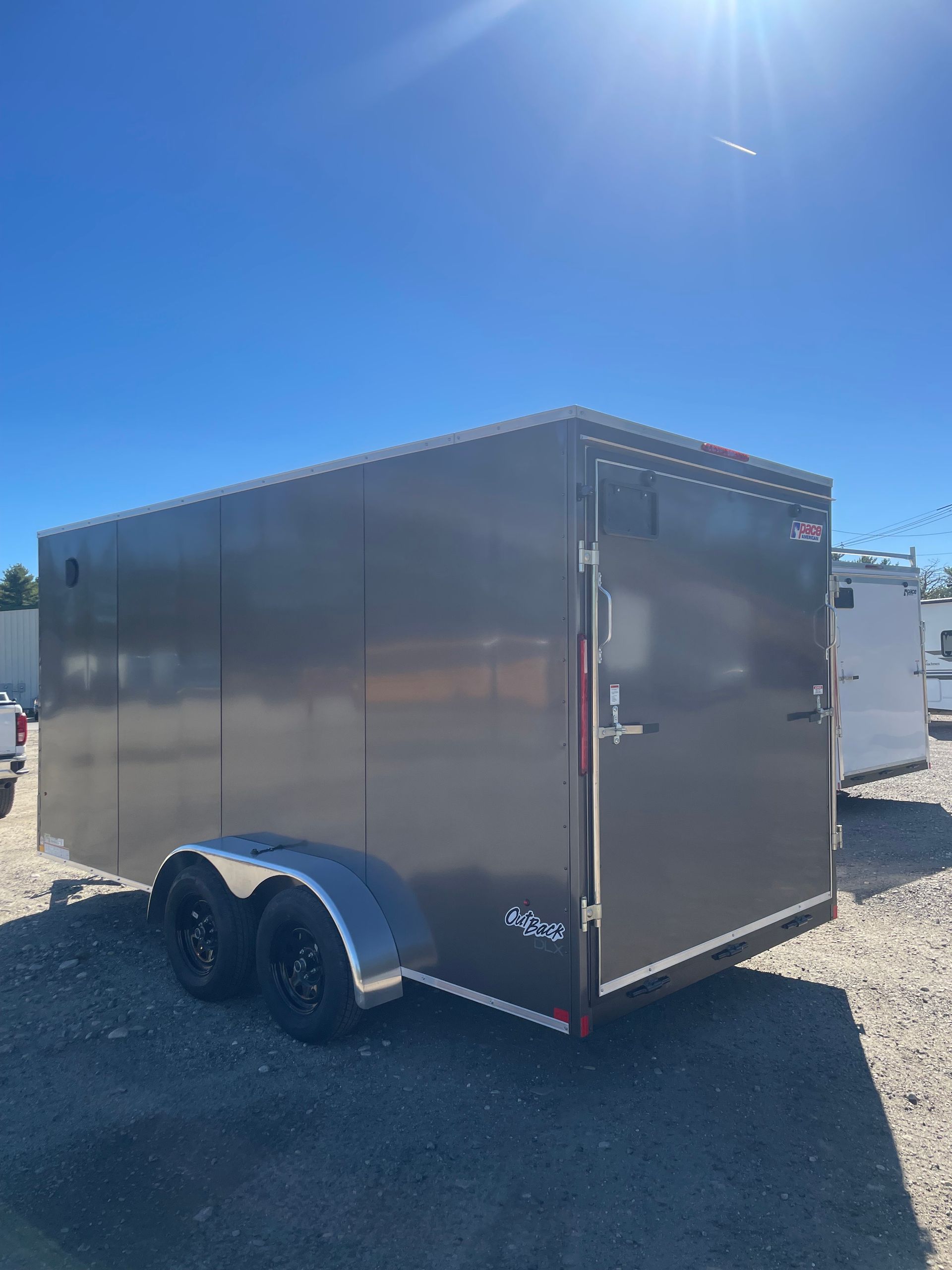 Gray enclosed trailer with black wheels parked on gravel under a bright blue sky.