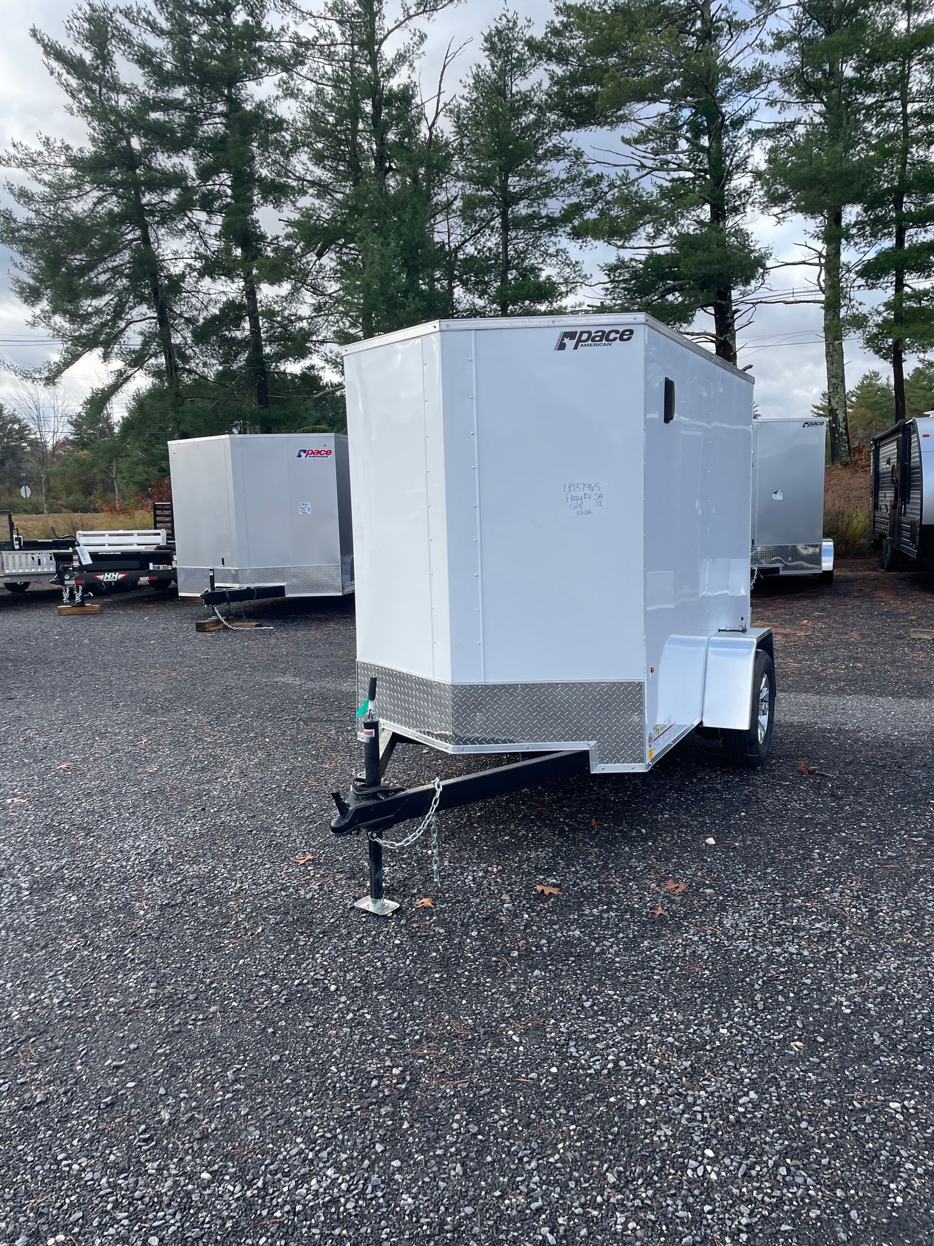 White enclosed utility trailer parked on a gravel lot; trees in the background.