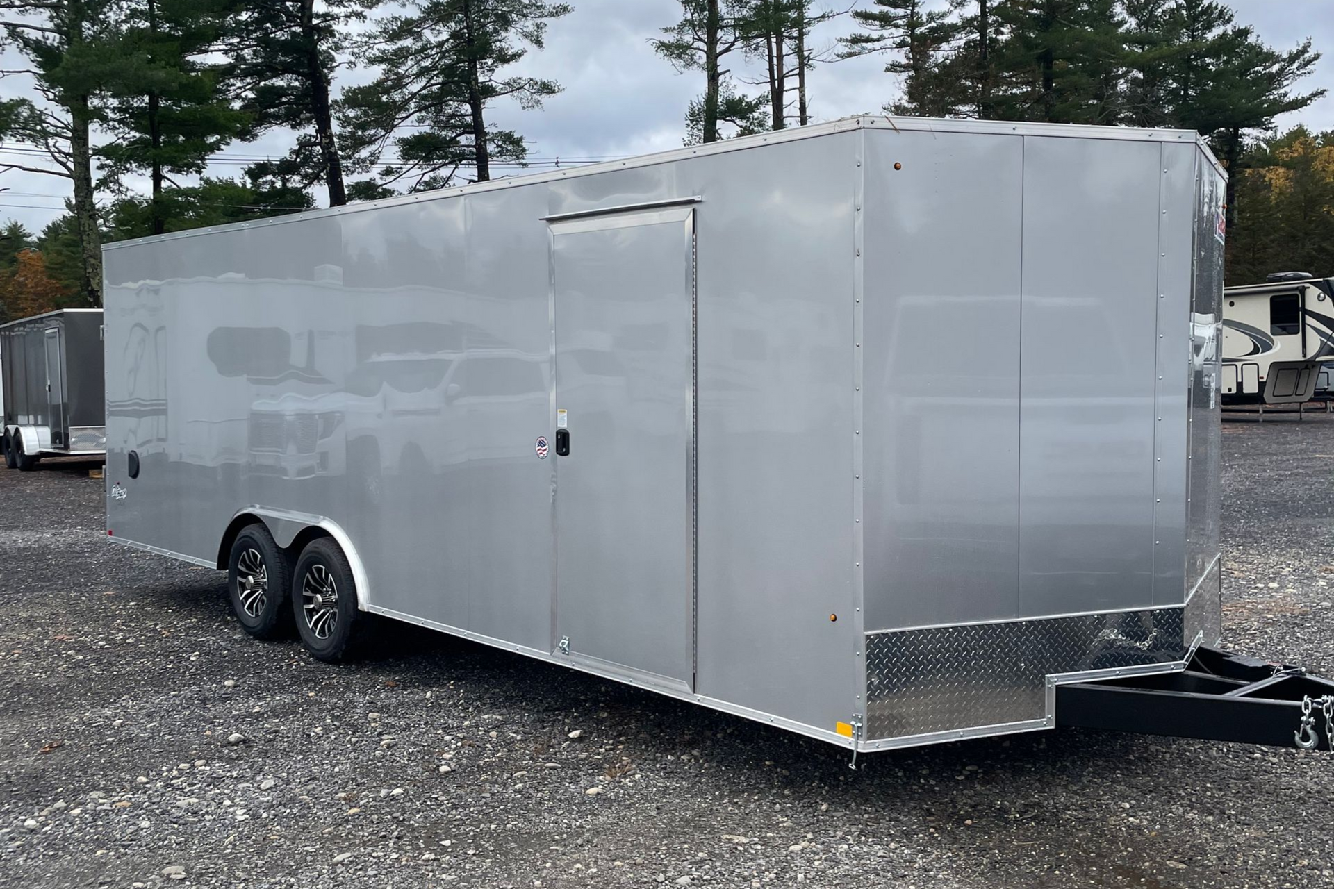 Silver enclosed trailer with a side door and black tires, parked outdoors on gravel.