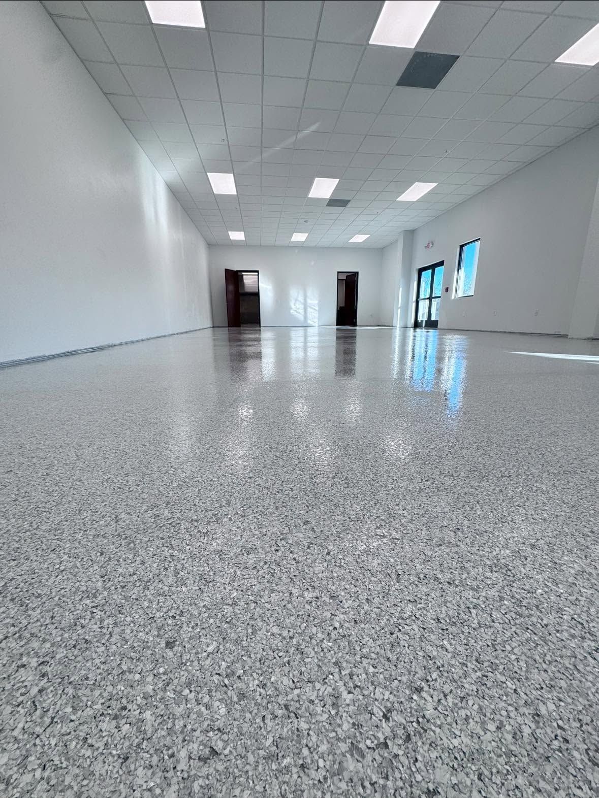 Empty white room with speckled floor, ceiling lights, and windows along the right wall