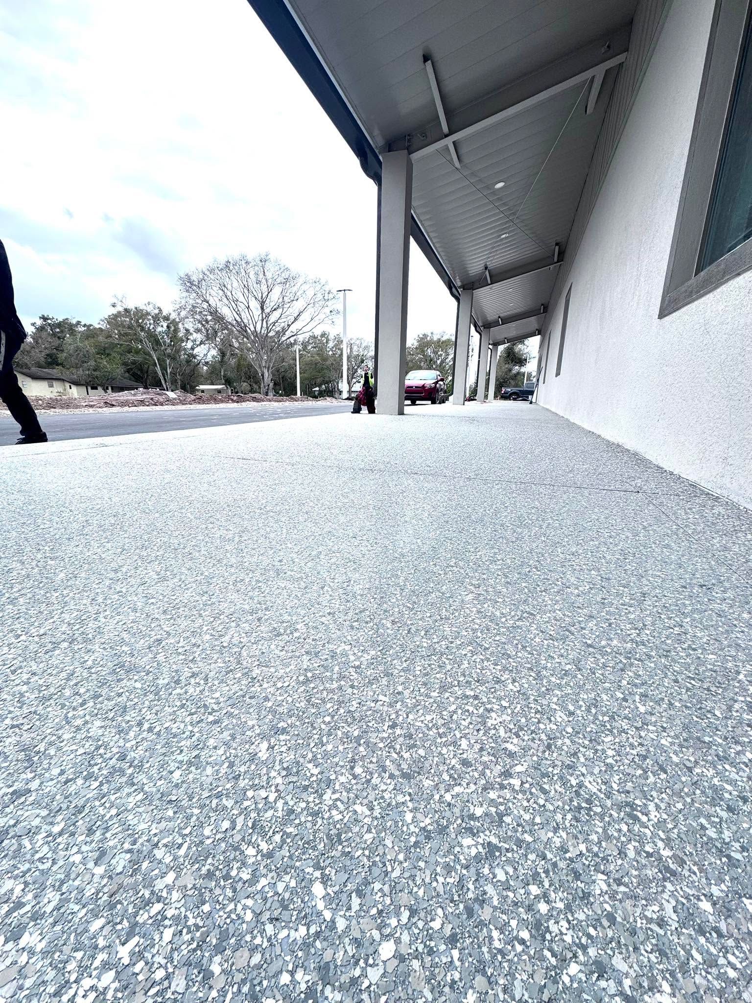 Low-angle view of a covered walkway with speckled floor, white wall, and trees in the distance.