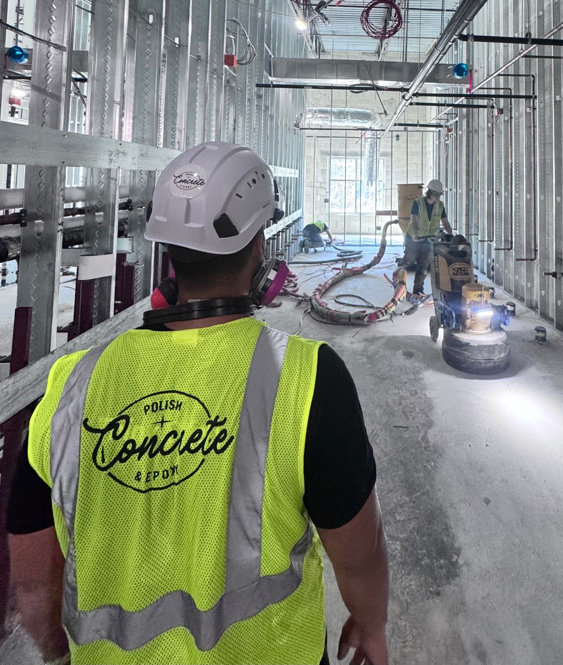 Worker in neon safety vest inside a construction site, walking toward a floor machine and exposed framing.